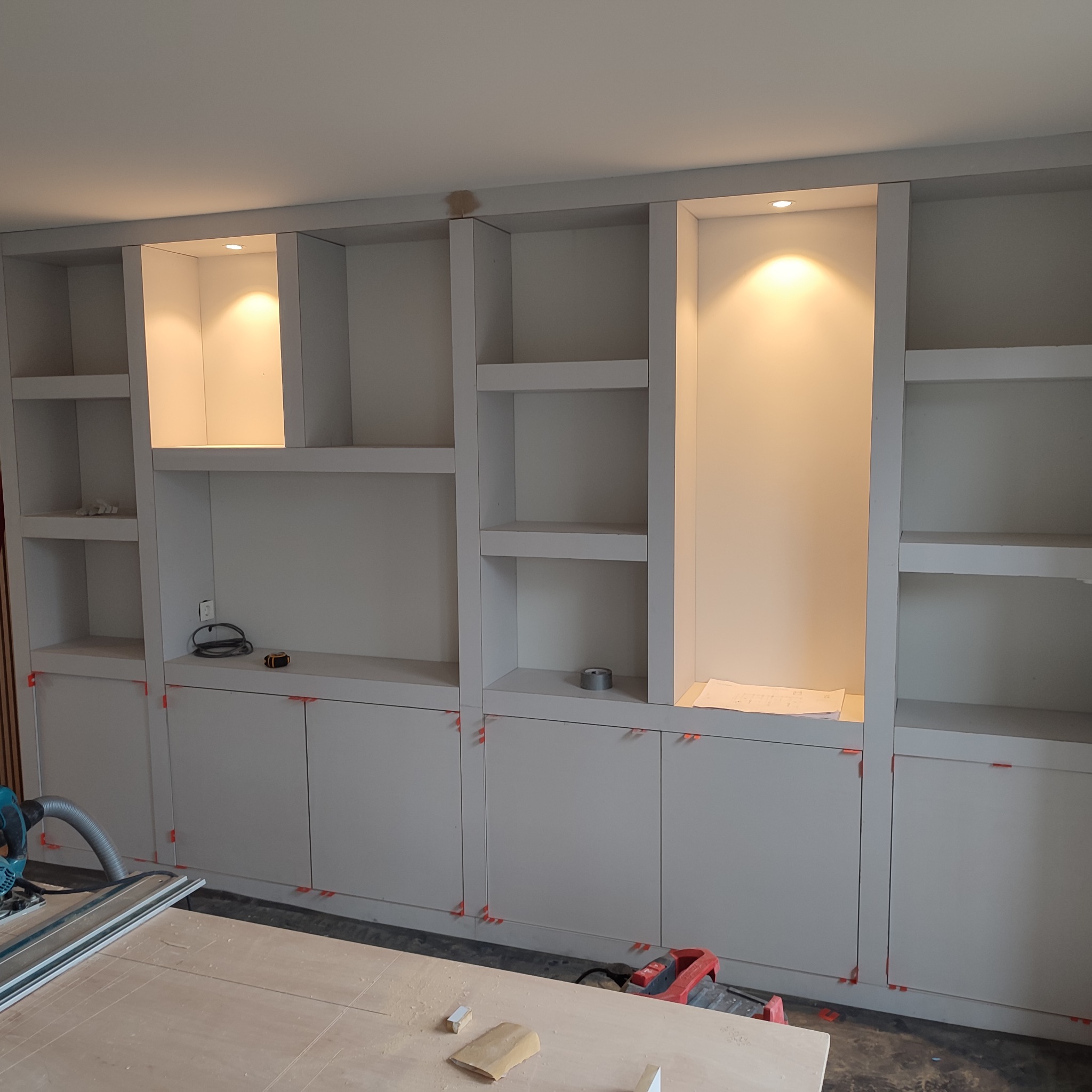 Nowoczesna, jasnoszara zabudowa meblowa z otwartymi półkami i szafkami, z oświetleniem punktowym w niektórych wnękach, widoczne narzędzia na blacie i podłodze, pomarańczowe kliny dystansowe przy...