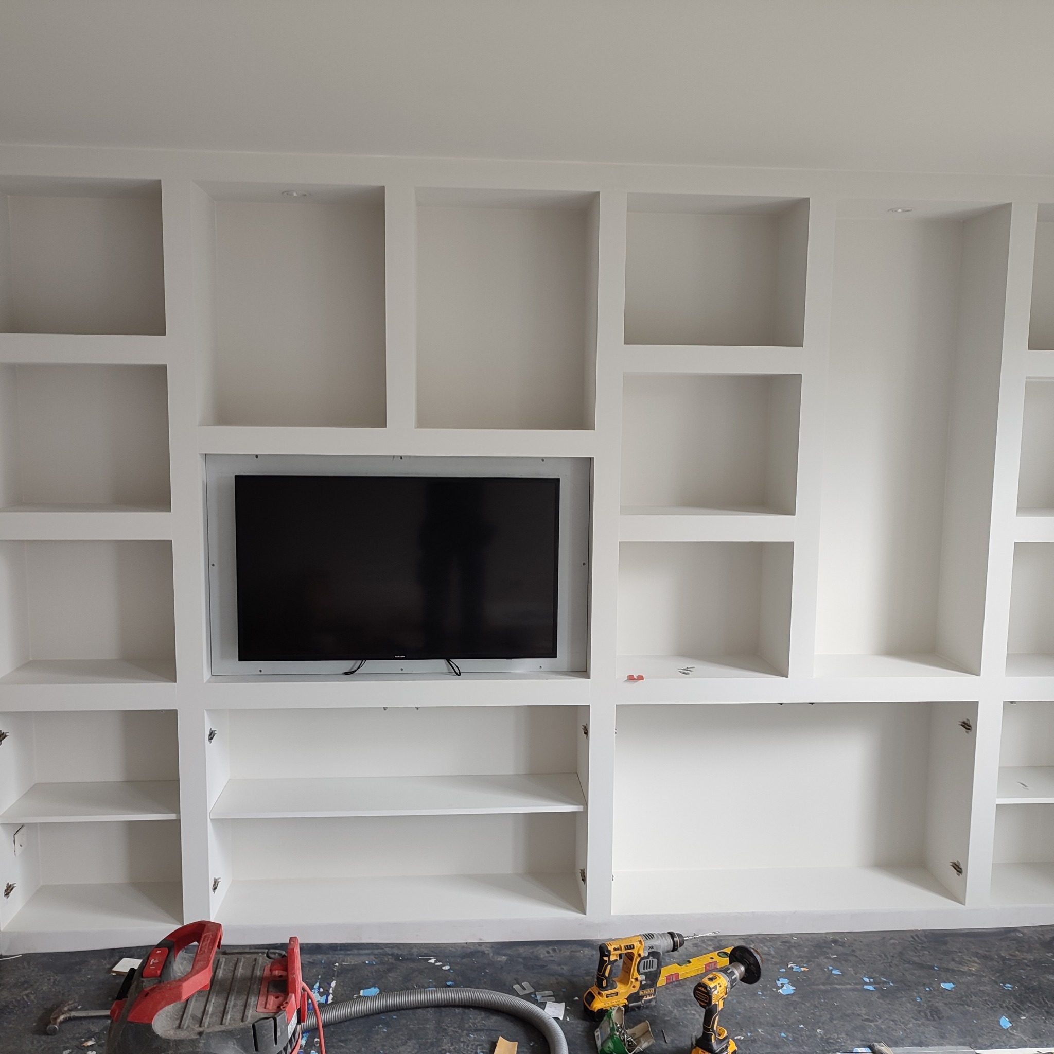 Biała zabudowa meblowa z wnęką na telewizor w trakcie montażu, widoczne narzędzia i odkurzacz na podłodze.
