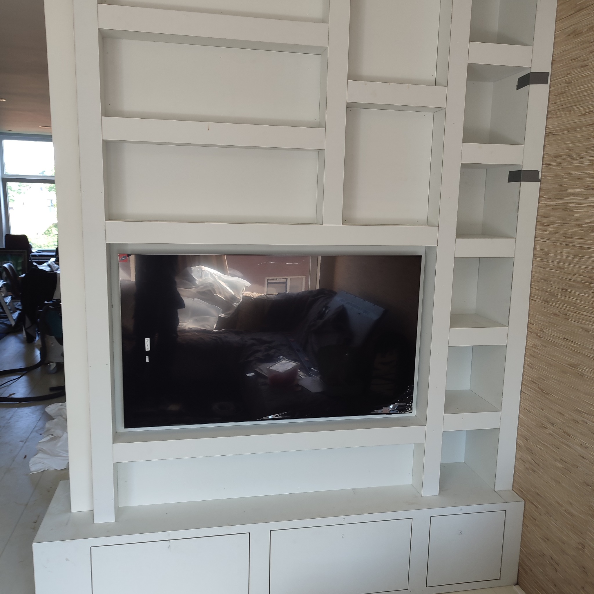 Biała zabudowa meblowa z wnęką na telewizor, widoczne półki i szafki, w tle bałagan remontowy.