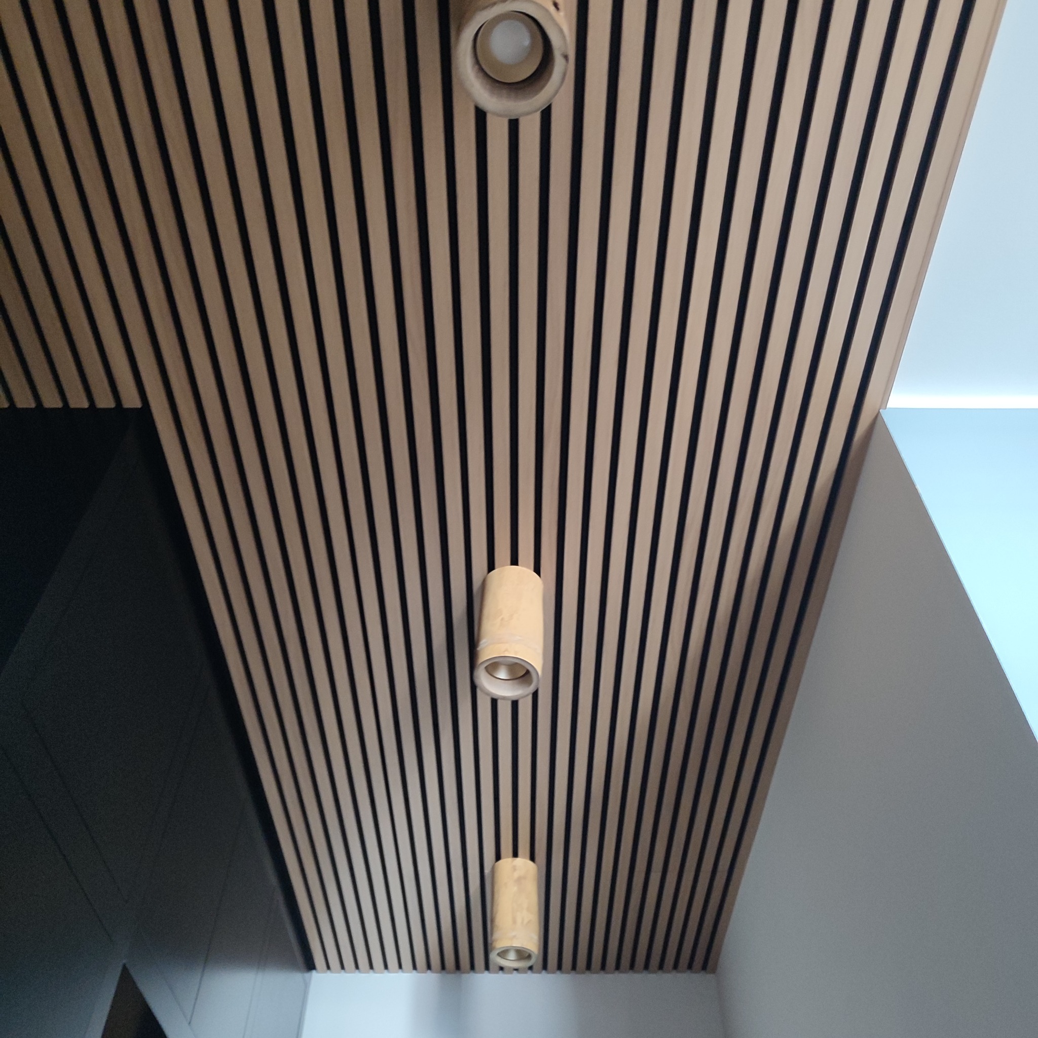 Drewniany sufit z równoległymi listwami i trzema cylindrycznymi lampami wpuszczanymi, widok z dołu.