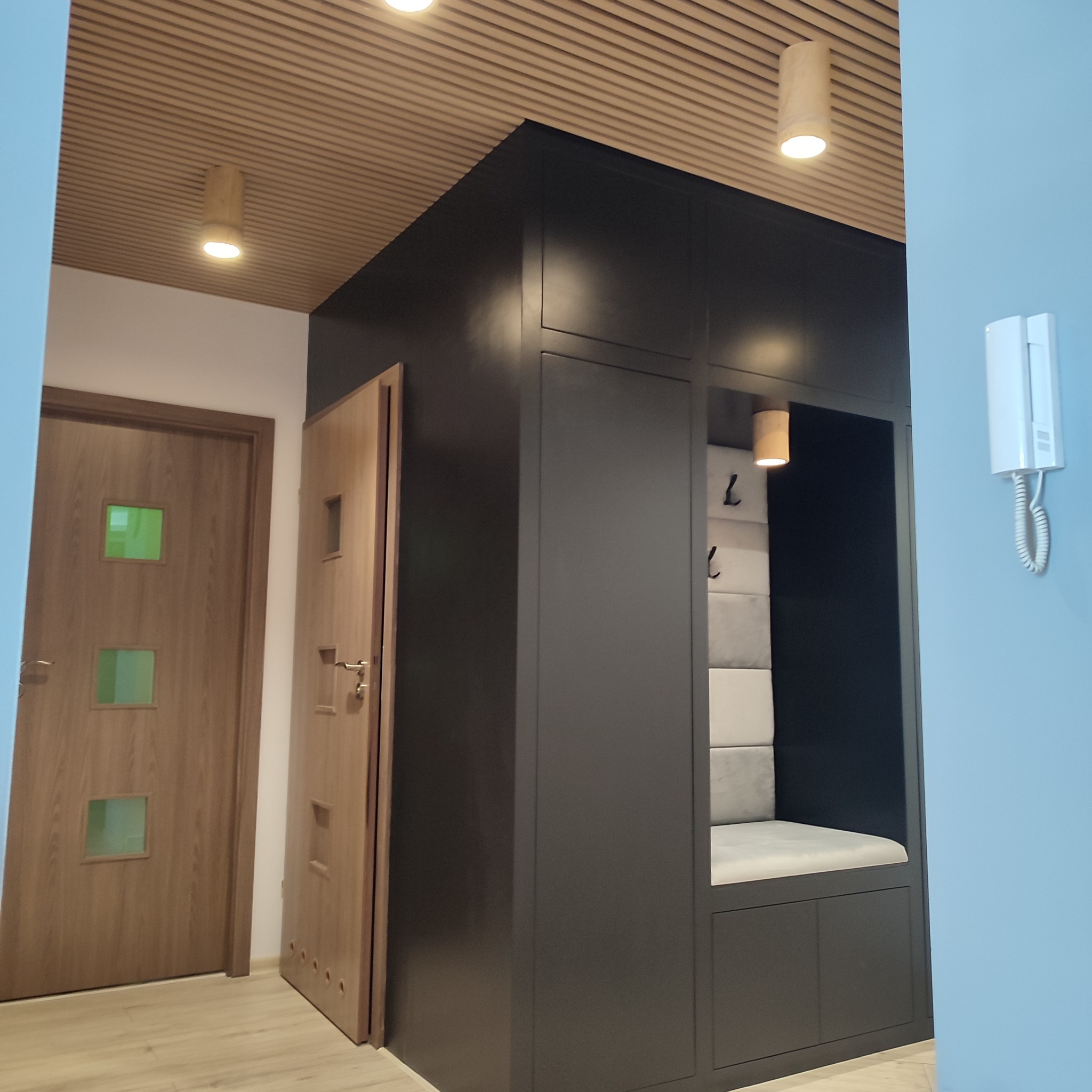 Nowoczesna zabudowa przedpokoju z siedziskiem i wieszakami, ciemna zabudowa meblowa, drewniany sufit z oświetleniem punktowym, widoczne drzwi wejściowe.