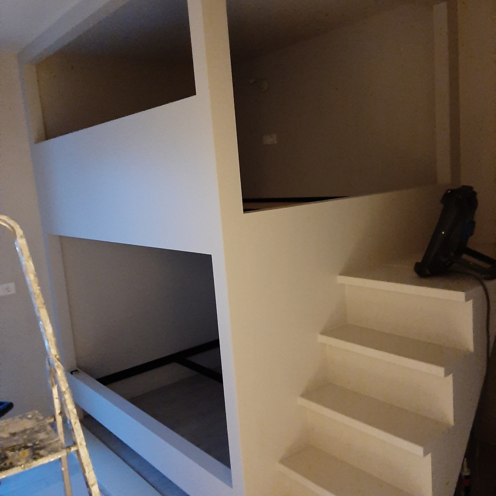 Białe, drewniane łóżko piętrowe z drabinką schodkową w trakcie montażu, widoczna drabina malarska i lampa halogenowa.