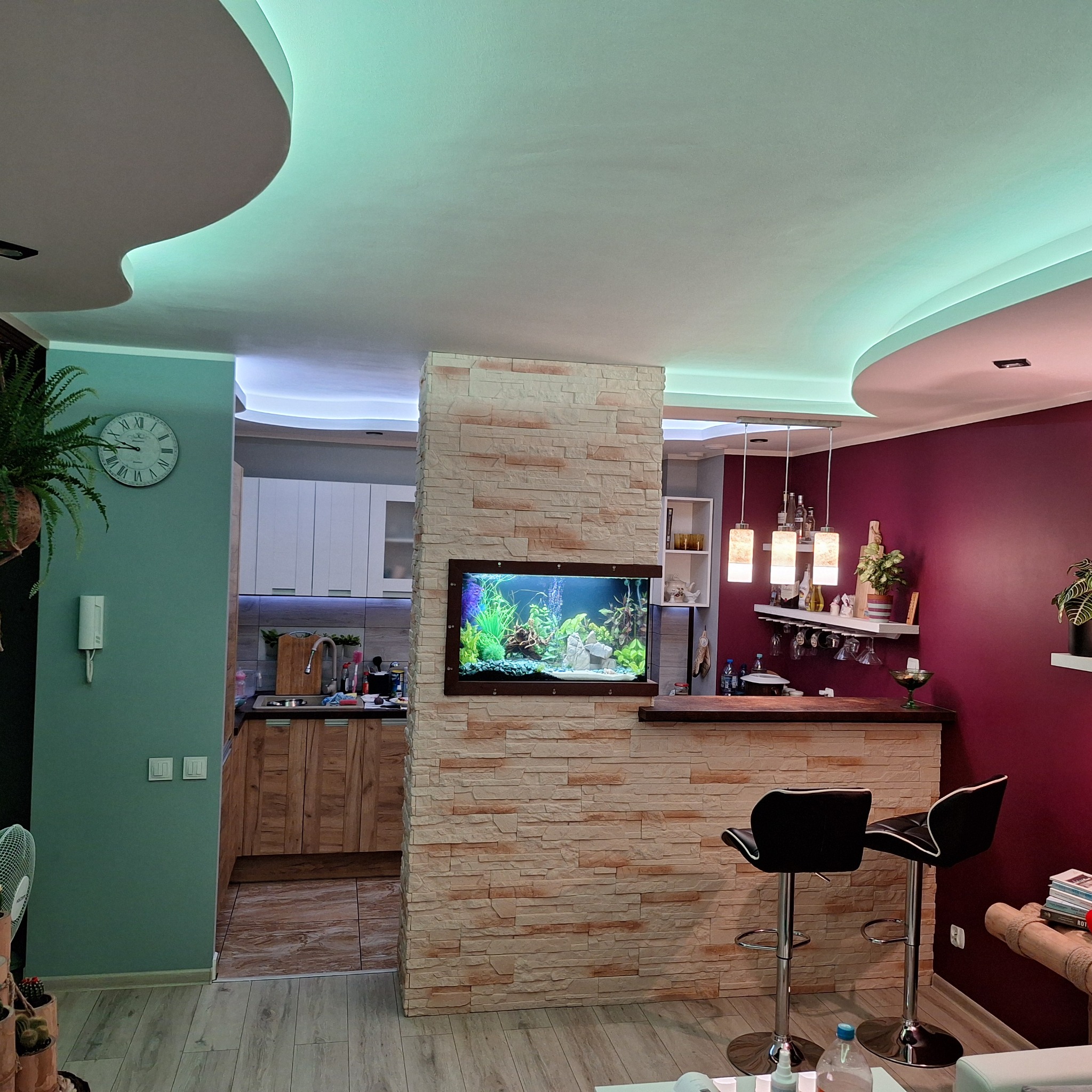 Nowoczesne wnętrze z akcentem w postaci akwarium wbudowanego w ścianę z imitacji kamienia, widoczna kuchnia w tle, oświetlenie LED pod sufitem i krzesła barowe.