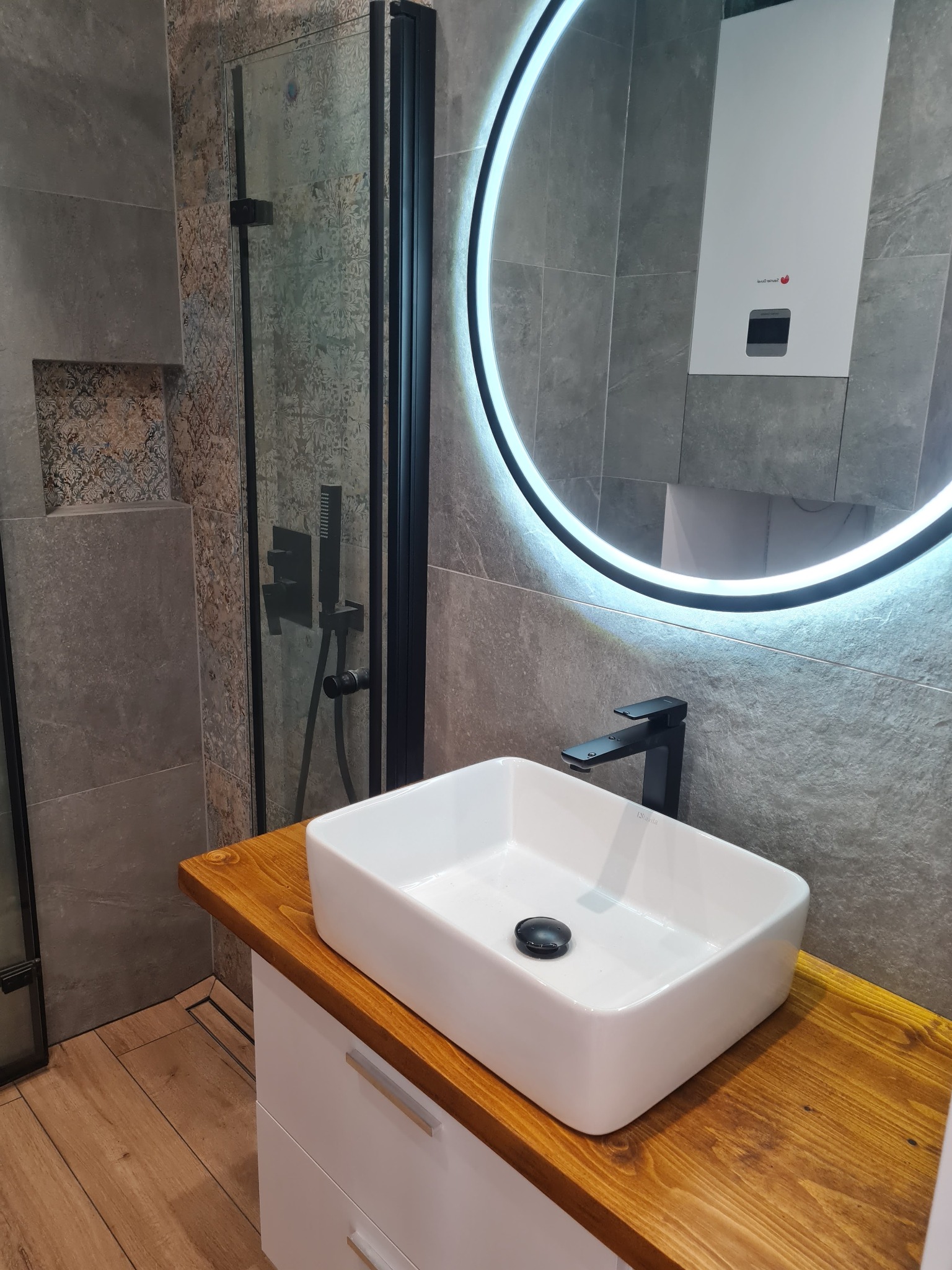 Nowoczesna łazienka z prostokątną białą umywalką nablatową, czarną armaturą i okrągłym lustrem z podświetleniem LED, szare płytki imitujące kamień na ścianach, kabina prysznicowa z czarnymi...