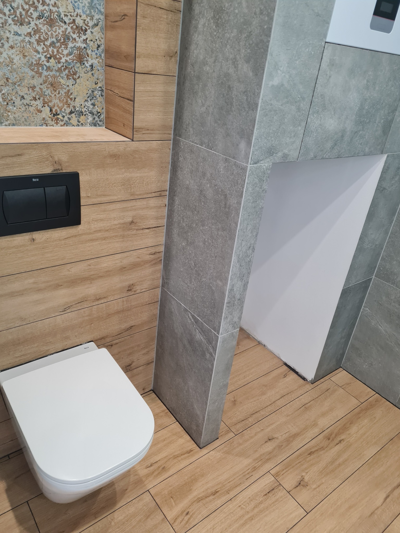 Nowoczesna łazienka z podwieszaną toaletą Roca, drewnopodobnymi płytkami na ścianach i podłodze oraz szarą, kamienną obudową stelaża.