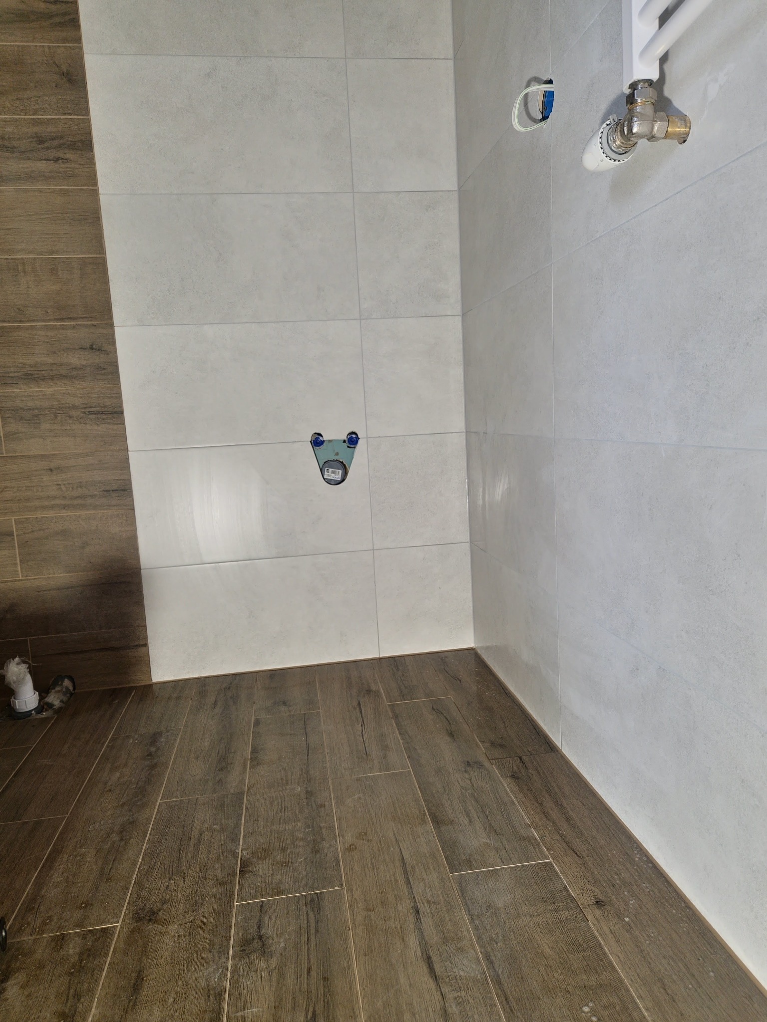 Wnętrze łazienki w trakcie remontu: szare płytki ścienne i brązowe płytki podłogowe imitujące drewno, widoczne przyłącza wodne i grzejnik, brakujące elementy wyposażenia.