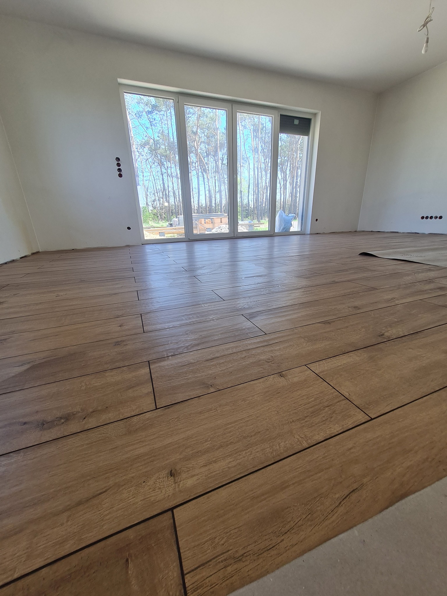 Ułożone panele podłogowe imitujące drewno w jasnym wnętrzu z dużym oknem balkonowym, widok na las sosnowy, widoczne niedokończone prace wykończeniowe przy ścianach.