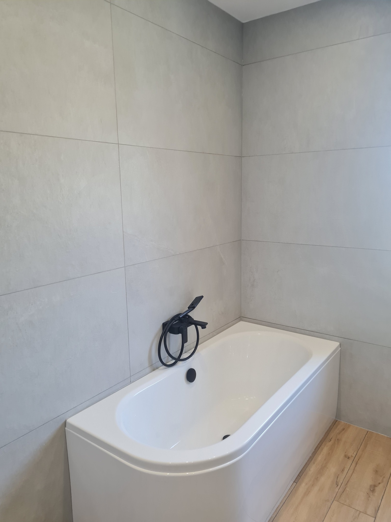 Wanna narożna z czarną armaturą na tle szarych płytek ściennych i podłogi imitującej drewno.