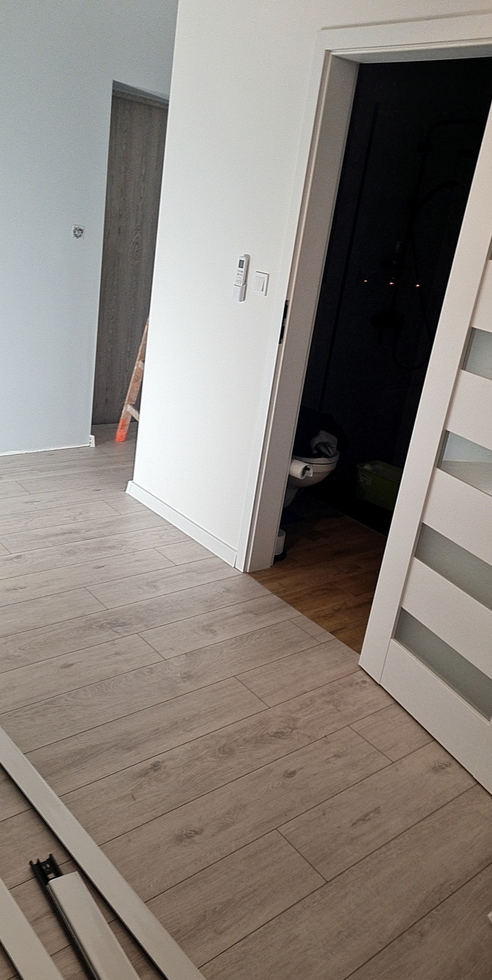 Jasne panele podłogowe ułożone w pokoju, widoczne listwy przypodłogowe, fragment drabiny malarskiej i toaleta w tle.