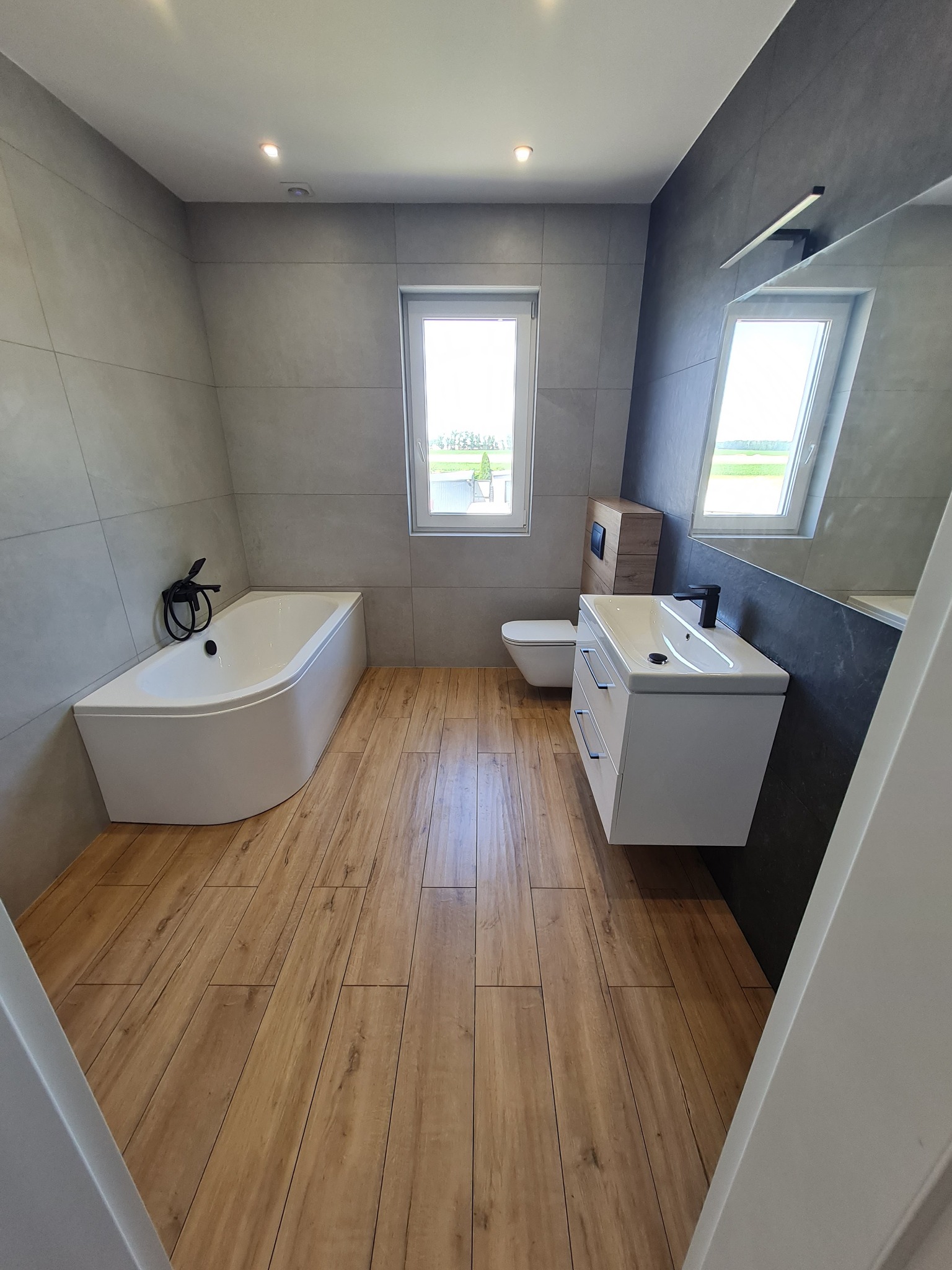 Nowoczesna łazienka z drewnopodobną podłogą, szarą glazurą ścienną, białą wanną narożną z czarną armaturą i wiszącą szafką umywalkową z czarną baterią, oświetlona punktowo.