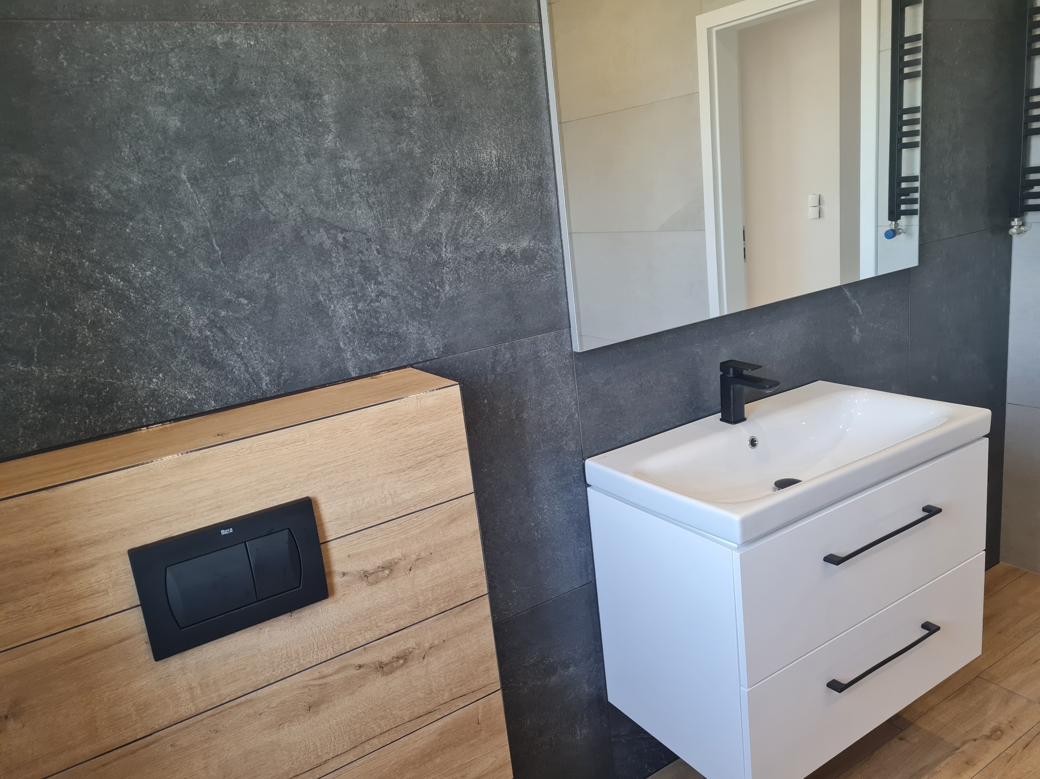 Nowoczesna łazienka z umywalką nablatową na białej szafce z czarnymi uchwytami, czarną armaturą i szarą płytką imitującą kamień na ścianie, oraz drewnopodobną zabudową stelaża WC z czarnym...