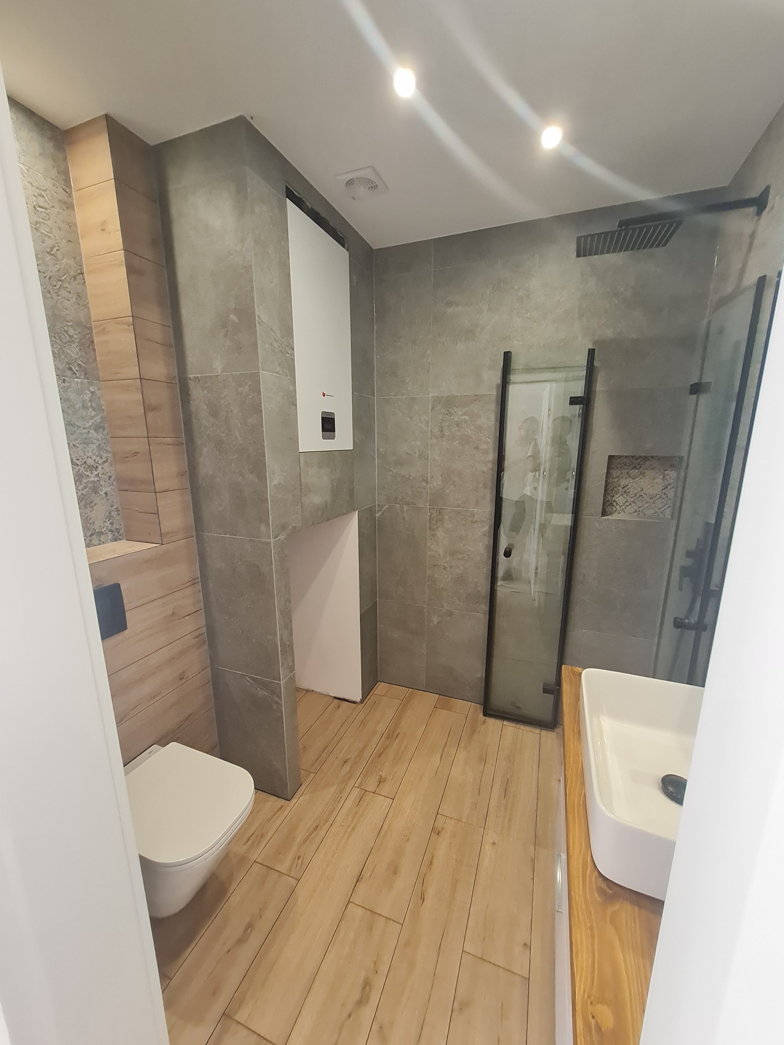 Nowoczesna łazienka z szarymi płytkami, drewnopodobnymi panelami na podłodze, kabiną prysznicową typu walk-in z czarnymi profilami, wiszącą toaletą i białą umywalką nablatową na drewnianym blacie.