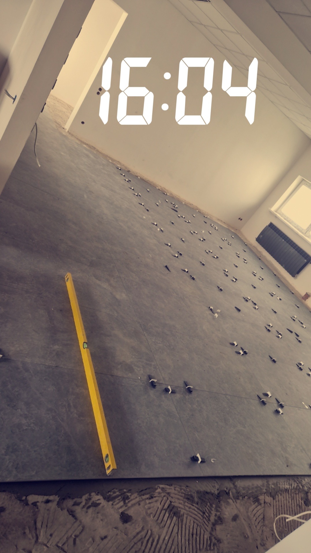 Układanie szarych płytek podłogowych z użyciem poziomicy i klinów dystansowych w jasnym pomieszczeniu, widoczny fragment grzejnika i okna, na zdjęciu nałożony filtr i cyfrowy zegar 16:04.