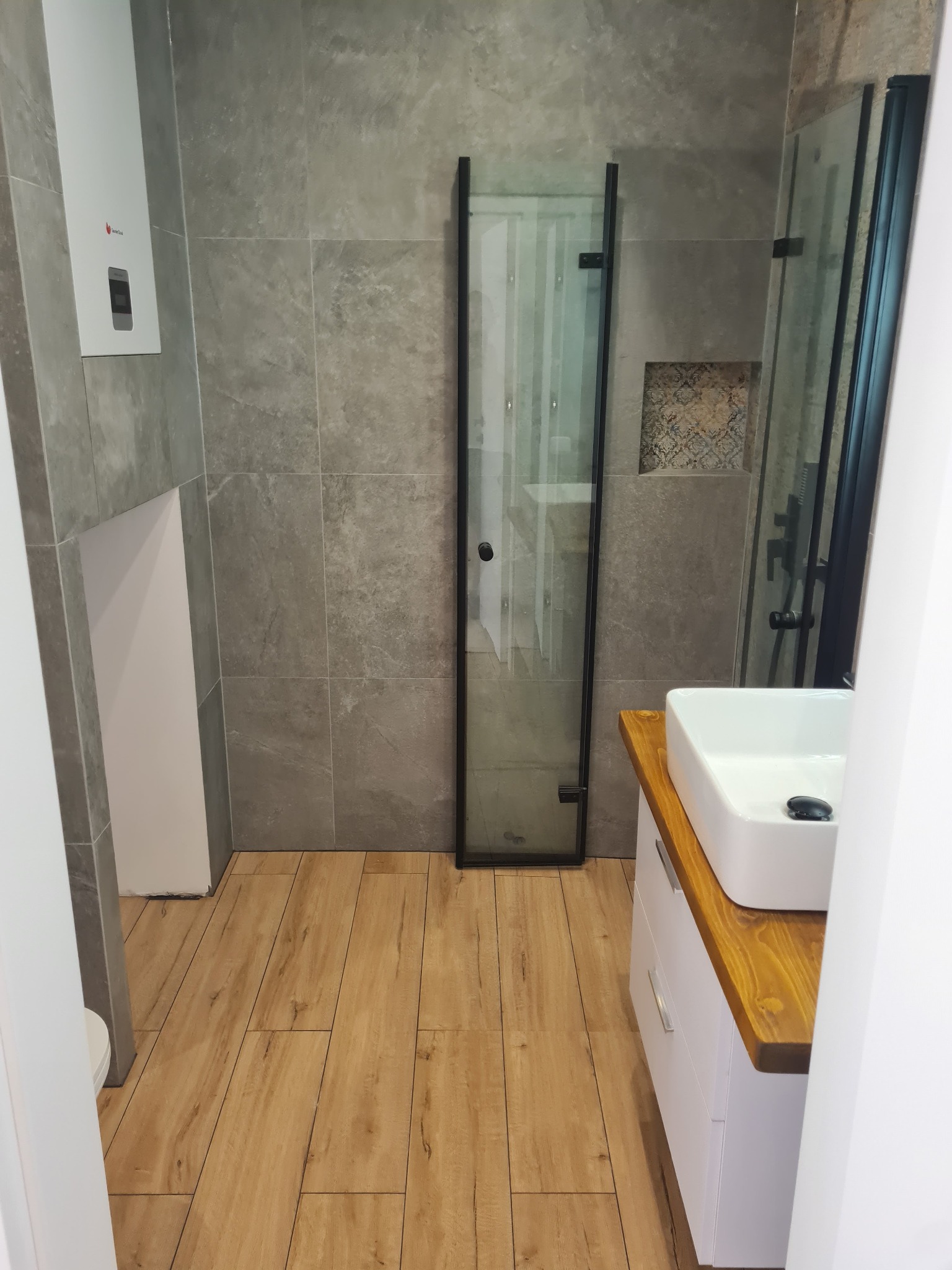 Wykończona łazienka w Buk z szarymi płytkami imitującymi kamień na ścianach, drewnopodobną podłogą, kabiną prysznicową z czarnymi ramami i białą umywalką nablatową na szafce z blatem z jasnego drewna.