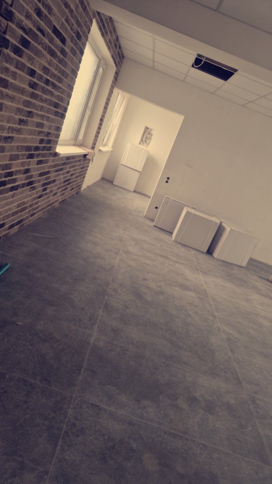 Wnętrze w trakcie wykańczania: ściana z cegły dekoracyjnej, szara podłoga z płytek imitujących kamień, białe elementy wyposażenia w tle, surowa ściana z widoczną instalacją elektryczną i otwór...