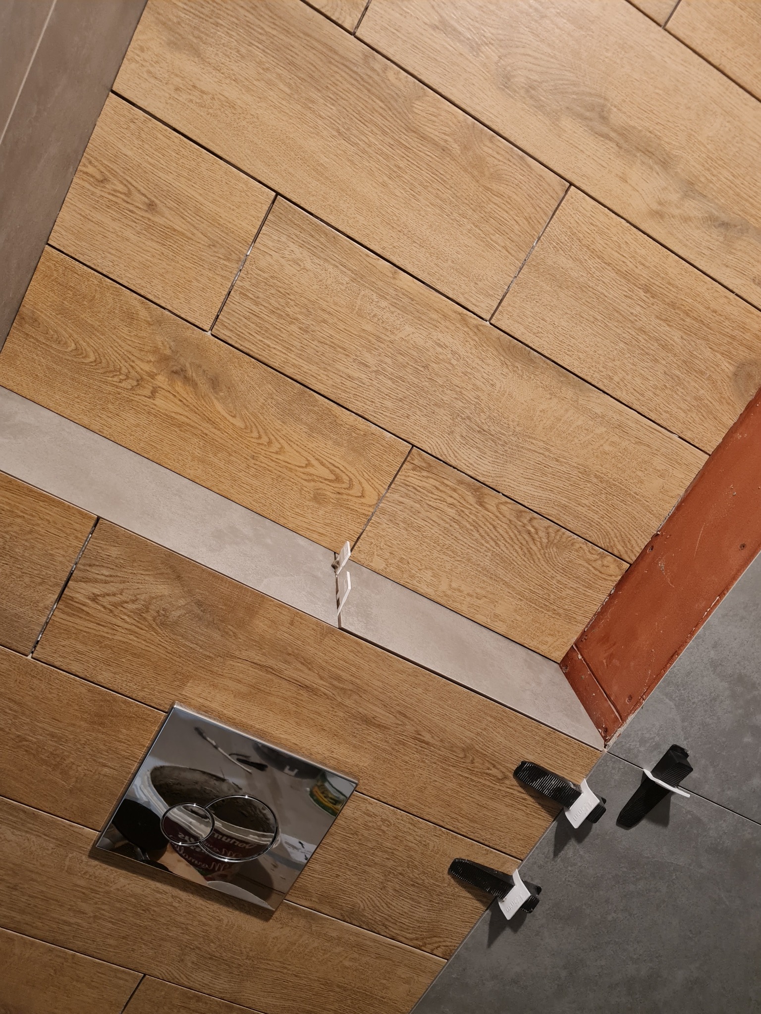 Układanie płytek imitujących drewno na podłodze w łazience, widoczne krzyżyki dystansowe, fragment ściany z czerwoną listwą oraz przycisk spłuczki WC.