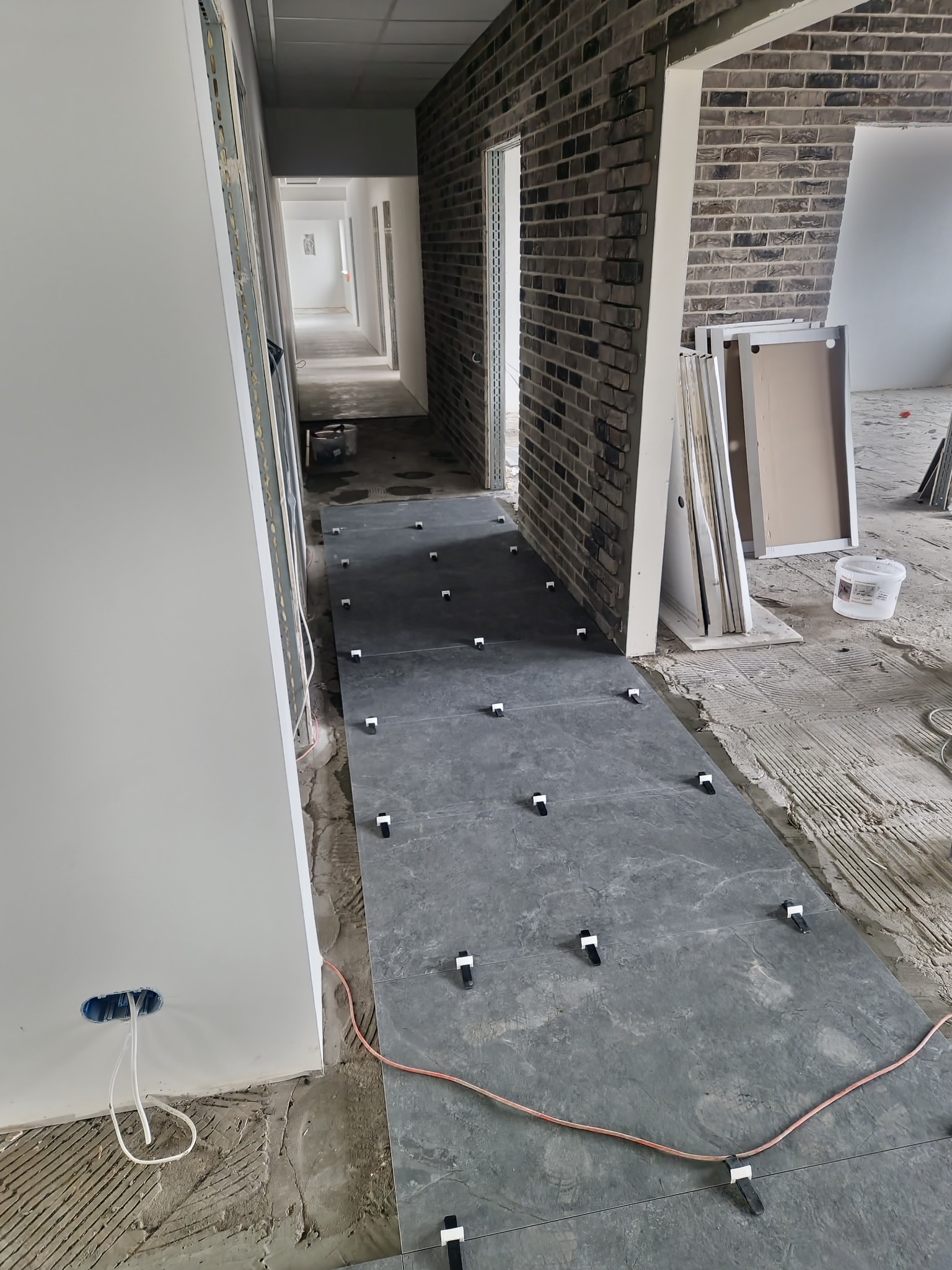 Kładzenie ciemnych płytek podłogowych w korytarzu, z użyciem klipsów poziomujących, widoczne przygotowane ściany z cegły i gładzi, instalacja elektryczna w ścianie.
