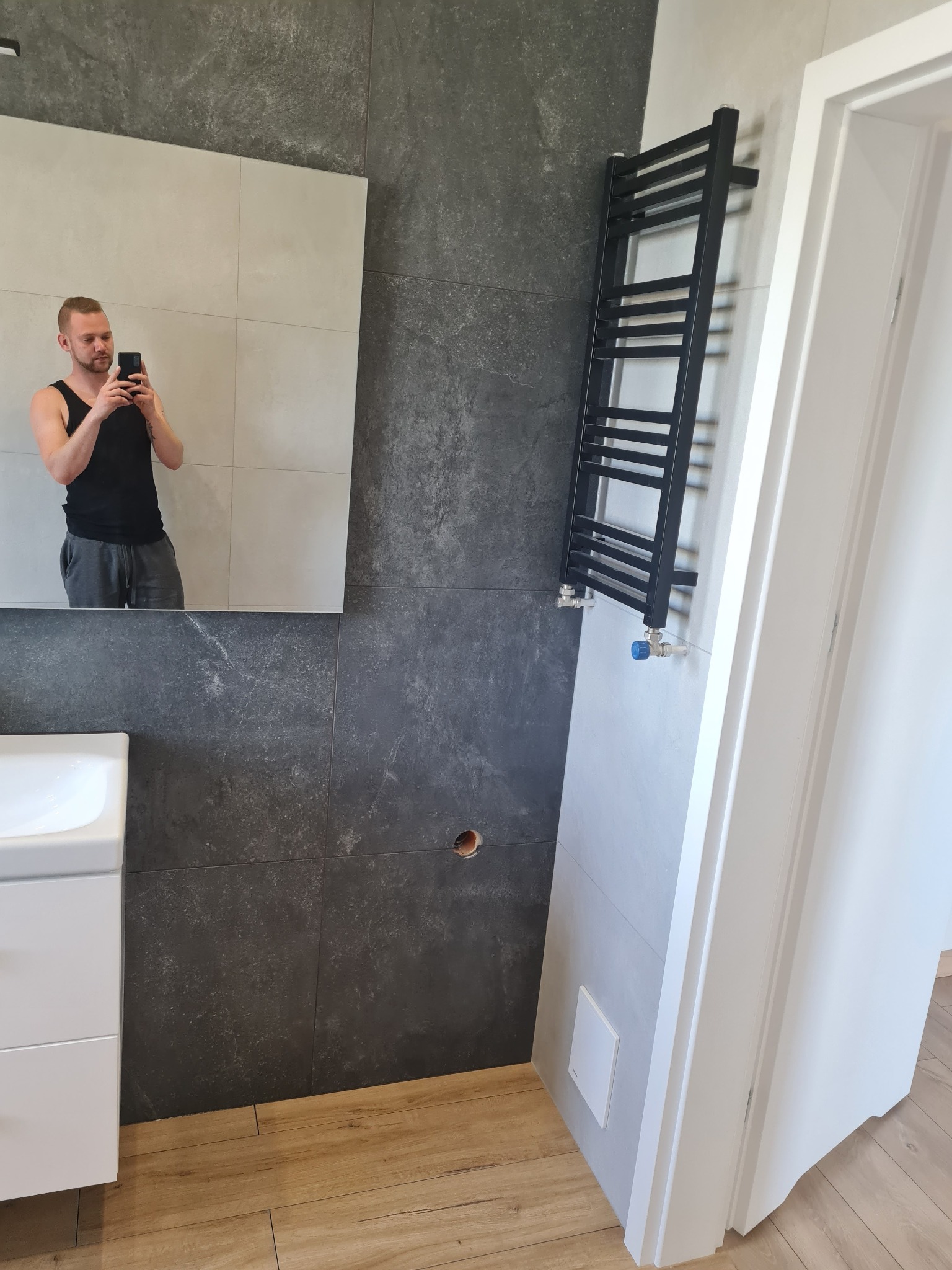 Ciemnoszara łazienka z drewnopodobną podłogą, widoczny grzejnik drabinkowy, umywalka z szafką i odbicie fotografa w lustrze.