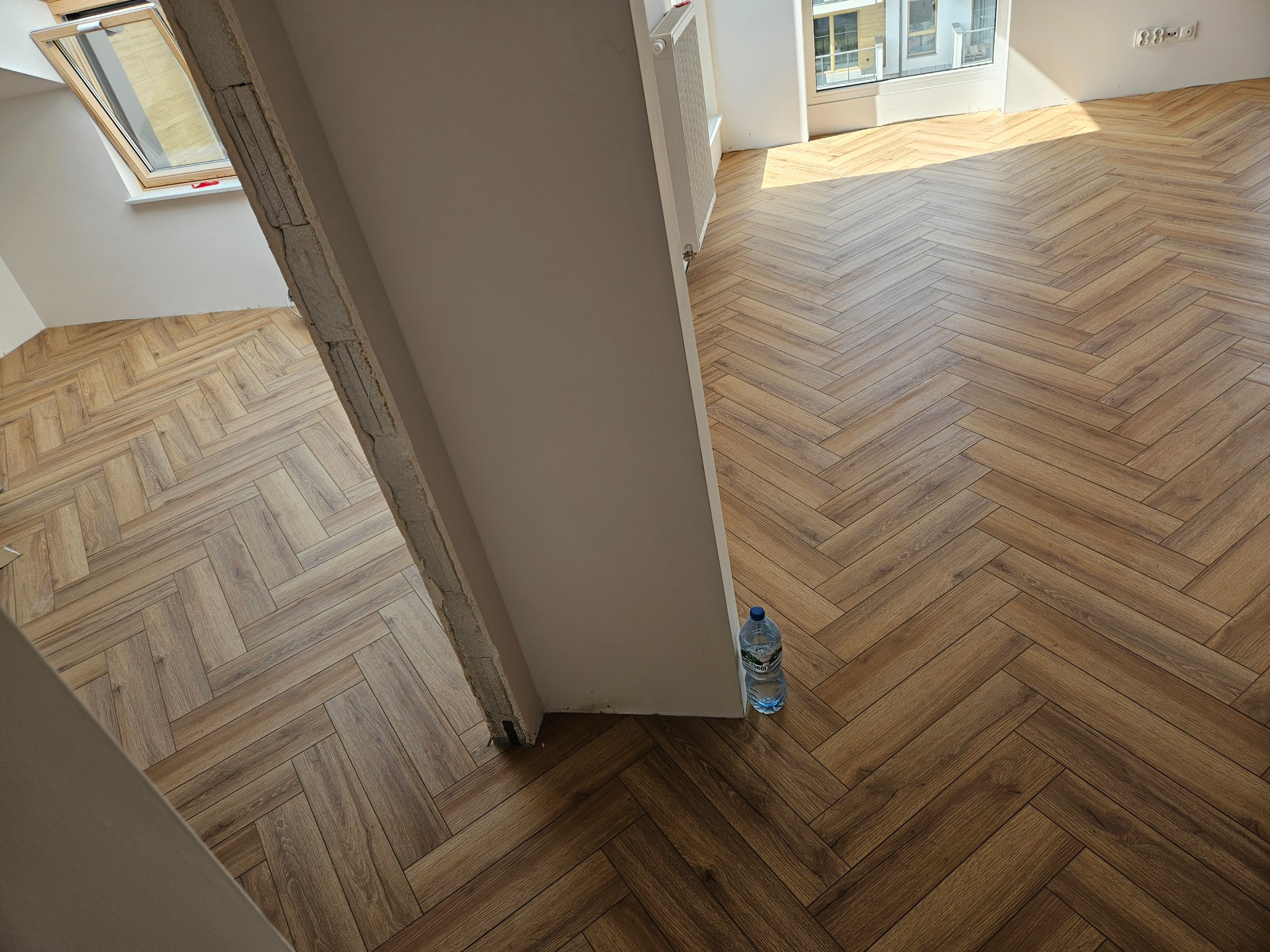 Ukończona podłoga z paneli ułożonych w jodełkę, widoczna przez otwarte drzwi, z butelką wody mineralnej przy ścianie.