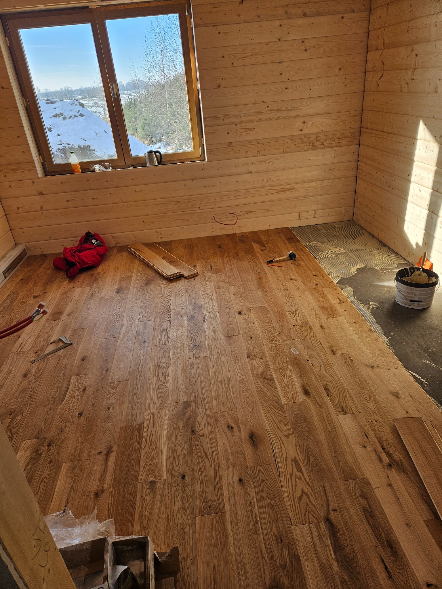 Fragment pomieszczenia z drewnianymi ścianami, w trakcie układania parkietu z widocznym klejem na podłożu, narzędziami i oknem z widokiem na zimowy krajobraz.