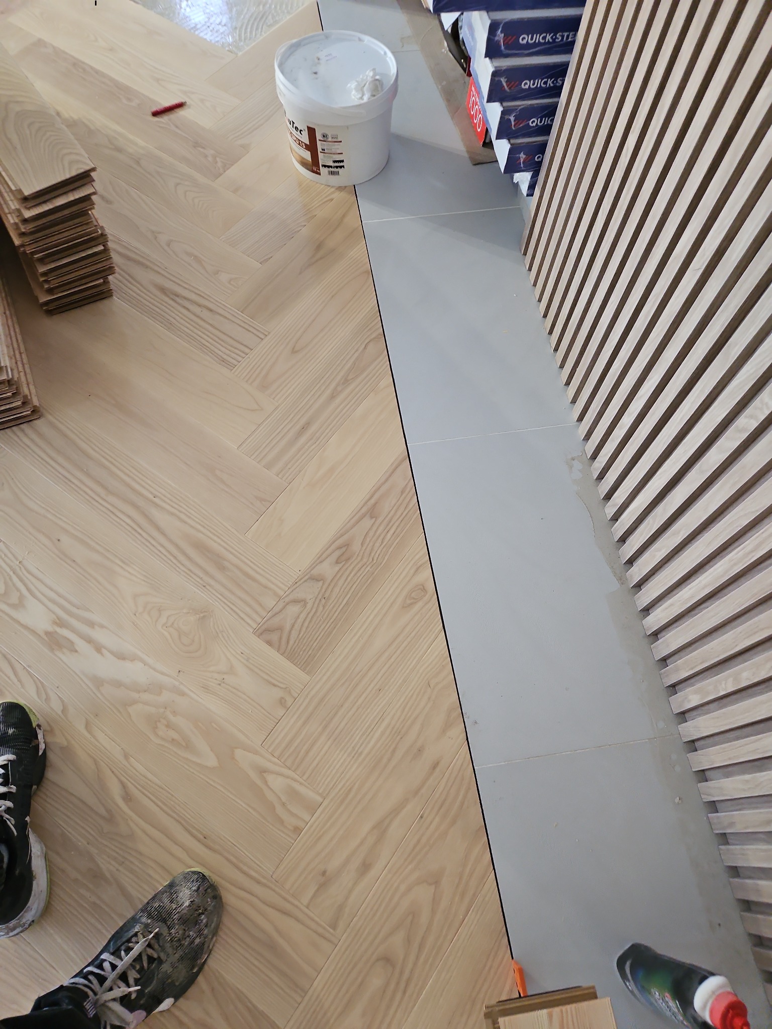 Jasny parkiet ułożony we wzór jodełki francuskiej, widoczny klej, paczki paneli, oraz listwy dekoracyjne na ścianie, buty robotnika w lewym dolnym rogu.