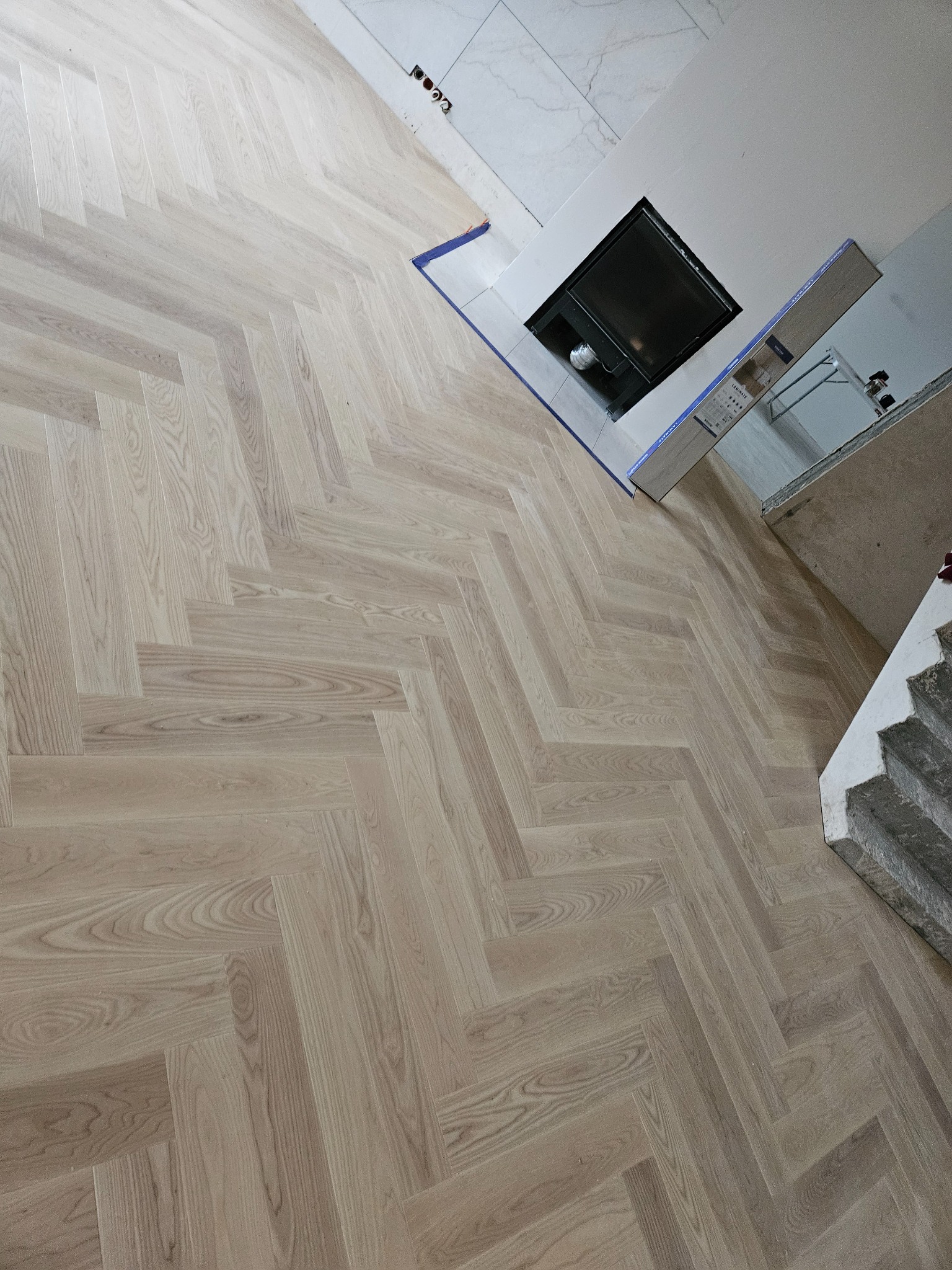 Ułożony parkiet w jodełkę francuską, jasne drewno, widoczne fragmenty ściany z gniazdkami elektrycznymi i kominek z wykończeniem, częściowo zakryty kartonami z panelami.