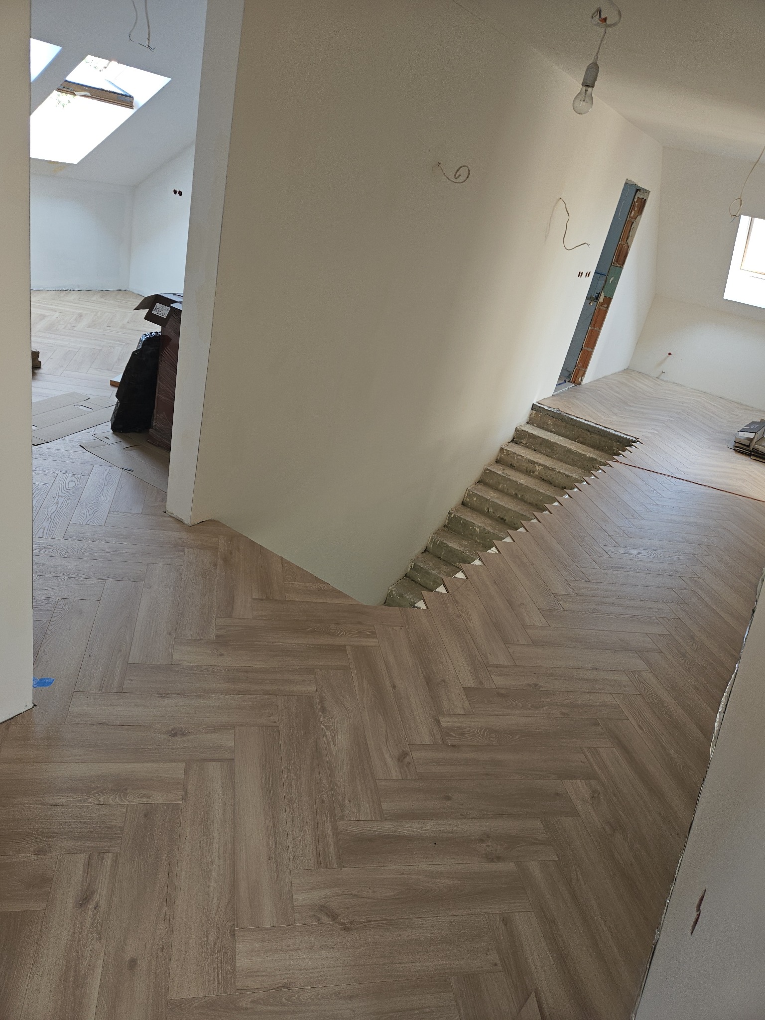 Ukończona podłoga z paneli ułożonych we wzór jodełki francuskiej, widok na schody w trakcie wykańczania i fragment poddasza z oknem dachowym w tle.