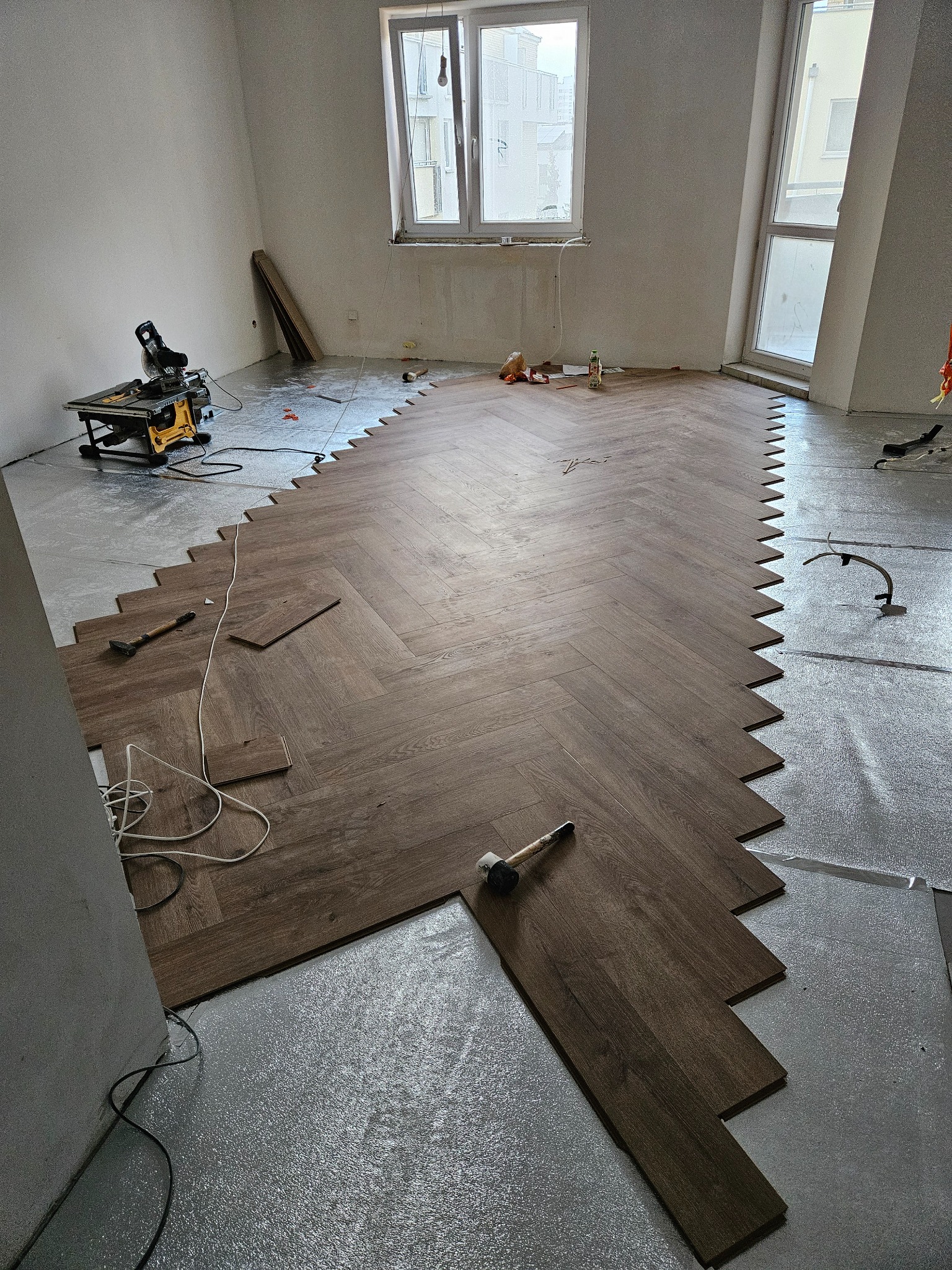 Układanie parkietu w jodełkę na podkładzie izolacyjnym, widoczna piła stołowa i gumowy młotek.