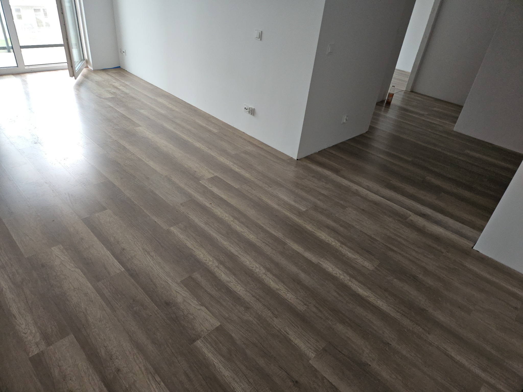 Ułożone panele podłogowe w odcieniach brązu z widocznym wzorem drewna, w jasnym, nowym mieszkaniu we Włocławku, z białymi ścianami i otwartym oknem balkonowym.