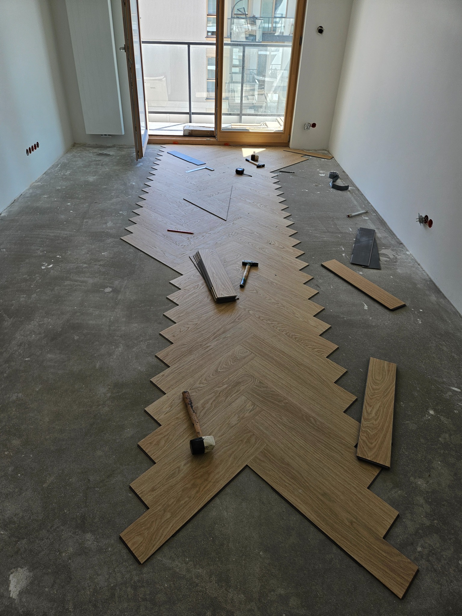 Układanie parkietu w jodełkę w jasnym pomieszczeniu z dużym oknem balkonowym. Na surowym podłożu widoczne narzędzia: gumowy młotek i dłuto.