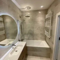 Jasna łazienka z beżowymi płytkami, okrągłe lustro z podświetleniem LED, biała umywalka z drewnianą szafką, wanna z parawanem, grzejnik drabinkowy.