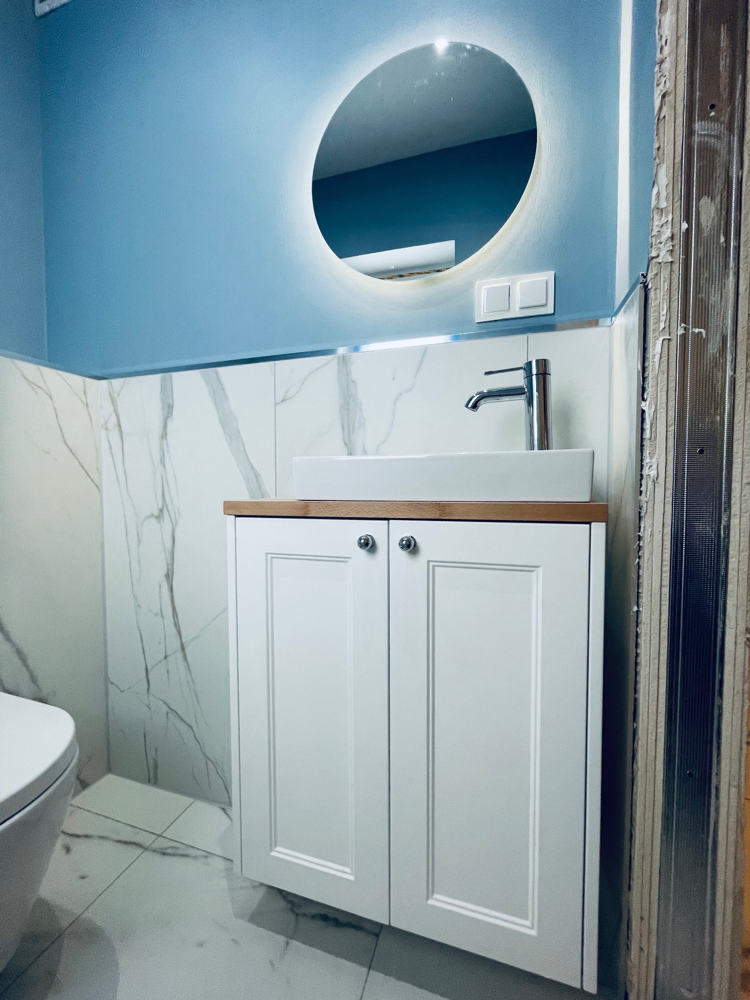 Nowoczesna łazienka w trakcie wykańczania: biała szafka pod umywalką z drewnianym blatem, okrągłe lustro z podświetleniem LED, marmurowe płytki na ścianach i podłodze, niebieska ściana, widoczne...