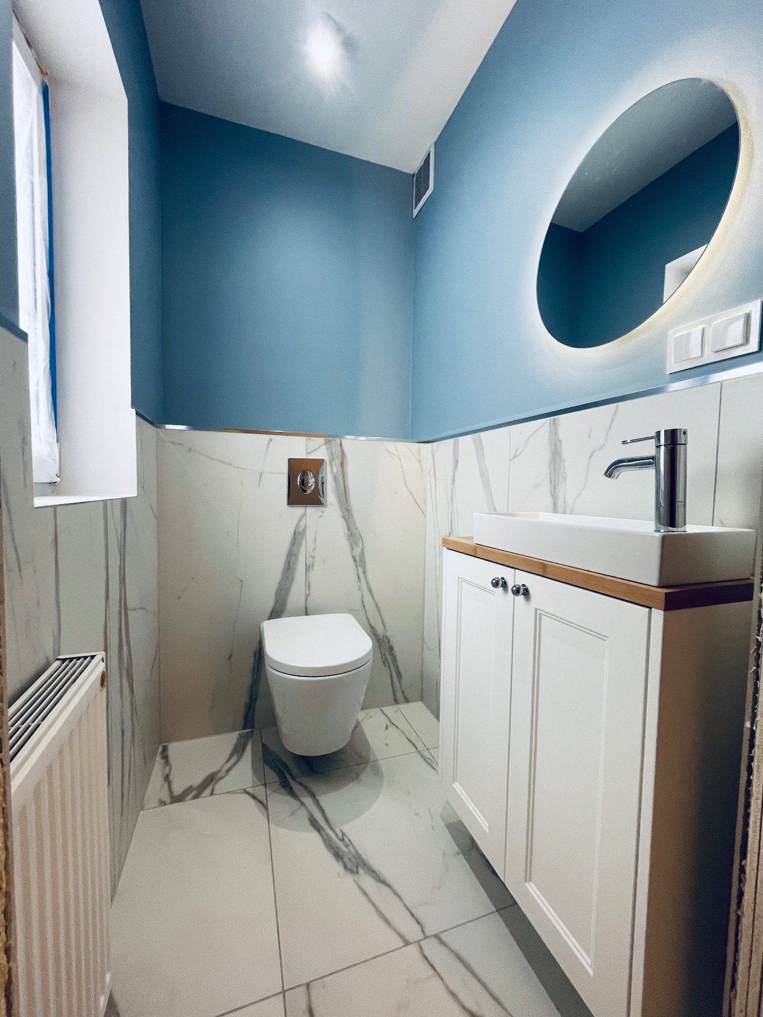 Nowoczesna łazienka w odcieniach błękitu i bieli z marmurowymi płytkami, podświetlanym lustrem owalnym, wiszącą toaletą i białą szafką pod umywalką.