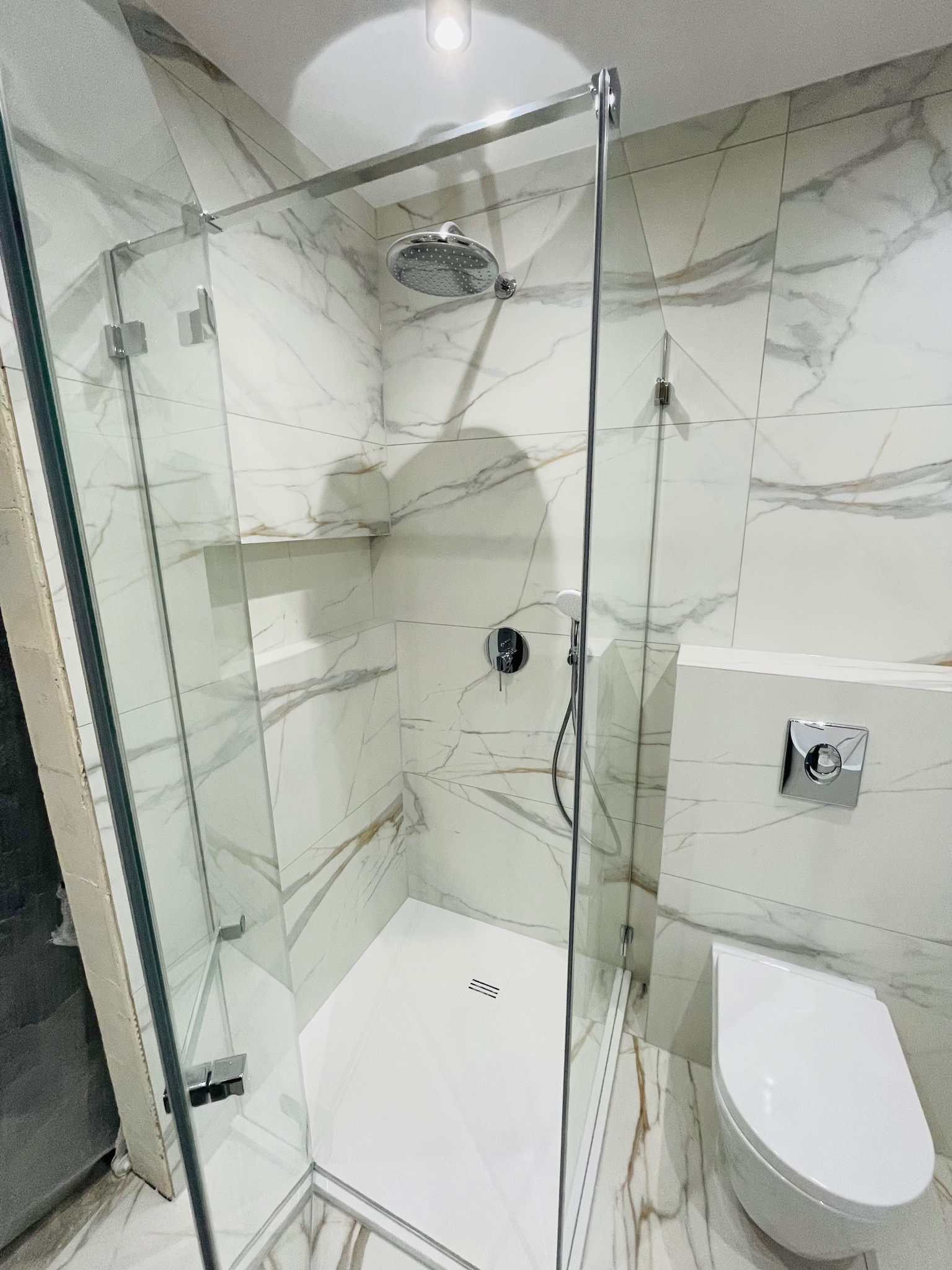 Nowoczesna kabina prysznicowa ze szklanymi drzwiami i deszczownicą, białe płytki imitujące marmur, wisząca toaleta z chromowanym przyciskiem spłukiwania, widoczny fragment surowej ściany przed...