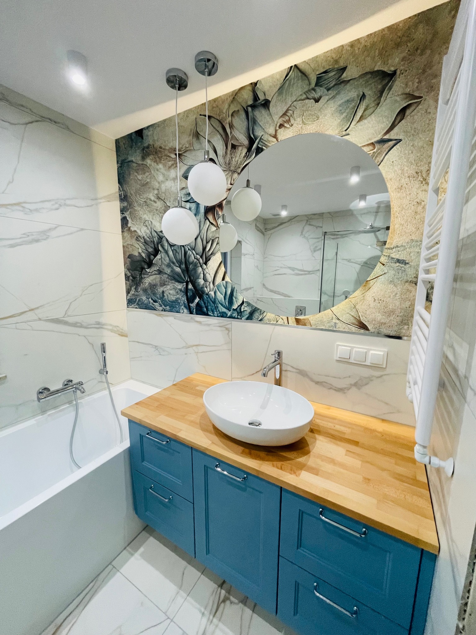 Nowoczesna łazienka z marmurowymi płytkami, okrągłym lustrem z motywem roślinnym, drewnianym blatem, niebieską szafką pod umywalką i grzejnikiem drabinkowym.