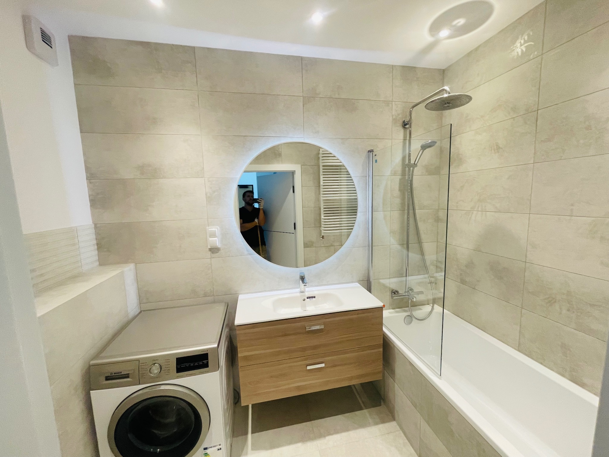 Nowoczesna łazienka z beżowymi płytkami, okrągłym lustrem z podświetleniem LED, białą umywalką z drewnianą szafką, pralką Bosch i wanną z parawanem nawannowym.