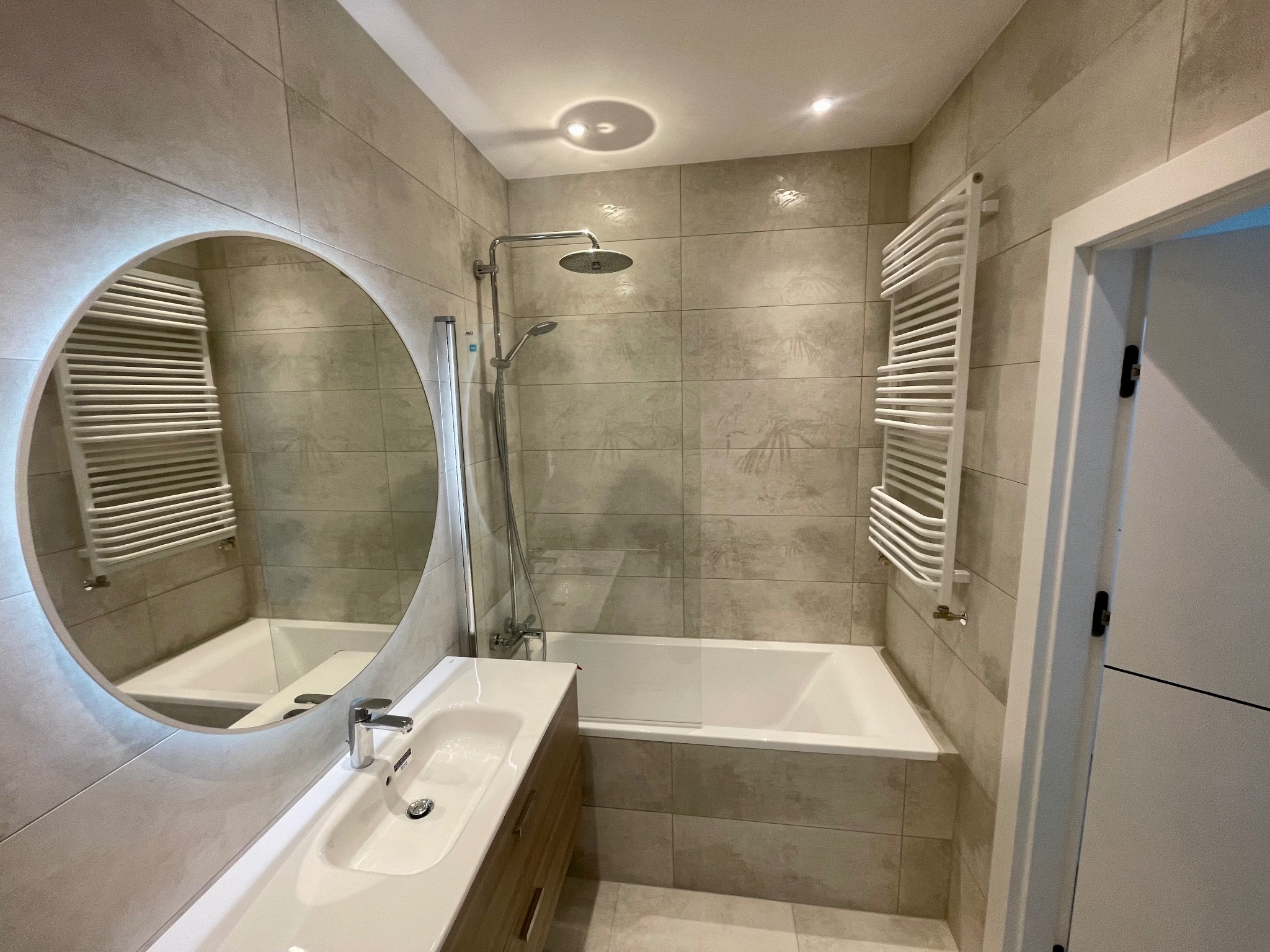 Jasna łazienka z beżowymi płytkami, okrągłe lustro z podświetleniem LED, biała umywalka z drewnianą szafką, wanna z parawanem, grzejnik drabinkowy.