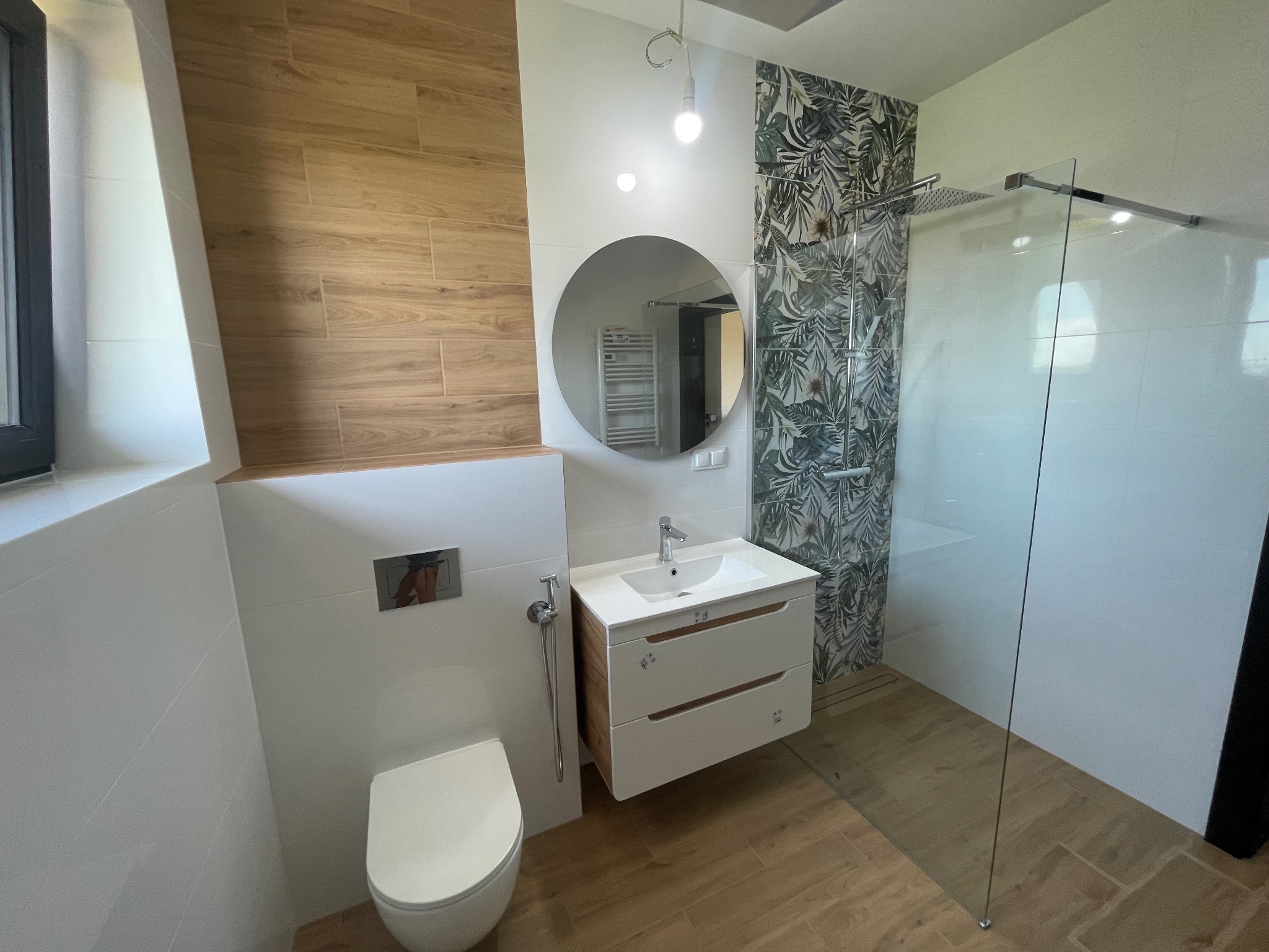 Nowoczesna łazienka z drewnopodobnymi płytkami, okrągłym lustrem i kabiną prysznicową typu walk-in z motywem roślinnym na ścianie.