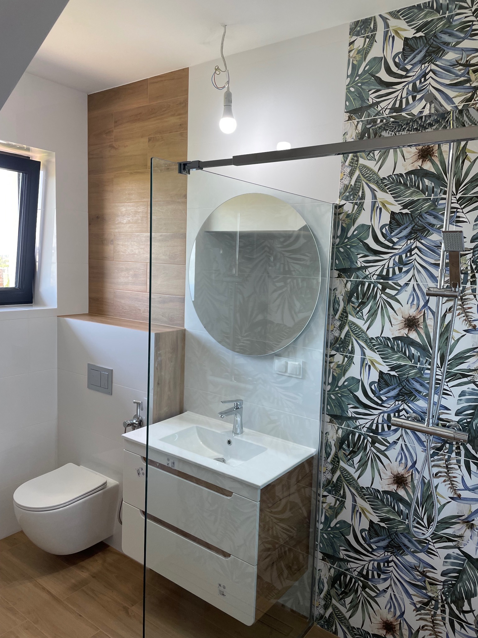 Nowoczesna łazienka z drewnopodobną podłogą i akcentem drewnianych płytek na ścianie, kabina prysznicowa ze wzorem tropikalnych liści, okrągłe lustro nad białą szafką z umywalką.