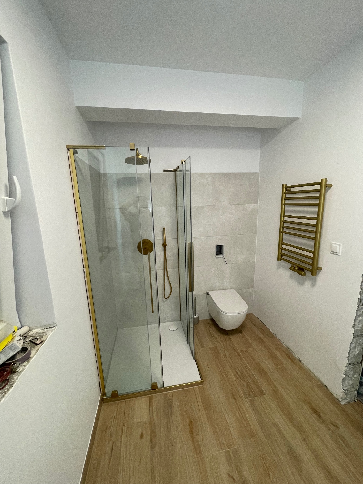 Nowoczesna łazienka z kabiną prysznicową ze złotymi ramami, wiszącą toaletą i grzejnikiem drabinkowym w kolorze złota, drewnopodobne płytki podłogowe, widoczne ślady prac wykończeniowych przy oknie...