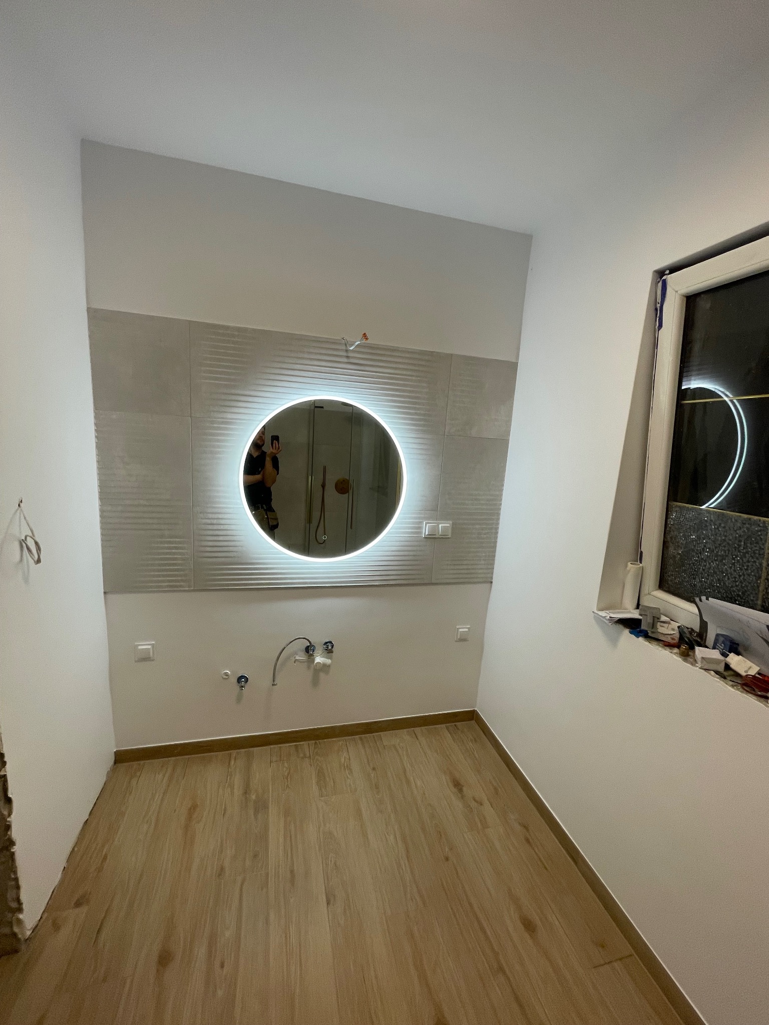 Łazienka w trakcie remontu z podświetlanym okrągłym lustrem, płytkami imitującymi fale na ścianie i drewnopodobną podłogą; widoczne niedokończone przyłącza wodne i okno z narzędziami na parapecie.