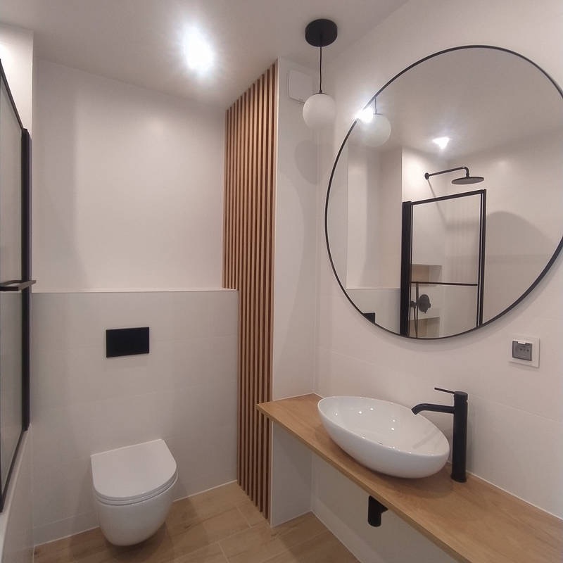 Nowoczesna łazienka z owalną umywalką nablatową, czarną armaturą i okrągłym lustrem w czarnej ramie. Ściana wykończona białymi płytkami, częściowo z dekoracyjnymi drewnianymi listwami. W tle...