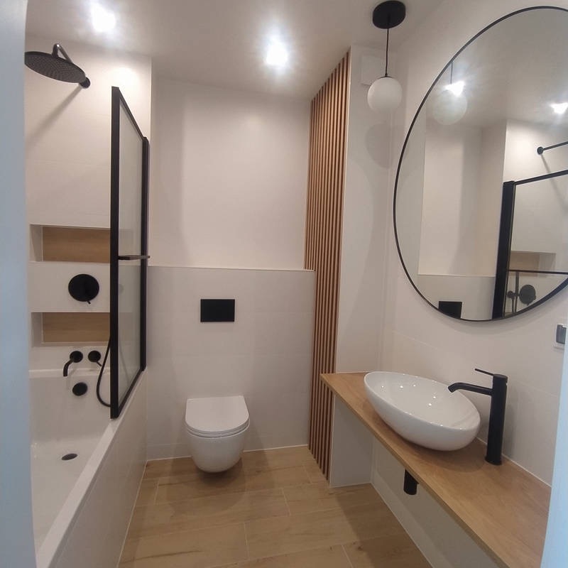 Nowoczesna łazienka z białymi płytkami, drewnianymi akcentami i czarną armaturą. Okrągłe lustro z czarną ramą nad umywalką nablatową. Kabina prysznicowa z czarnymi profilami.