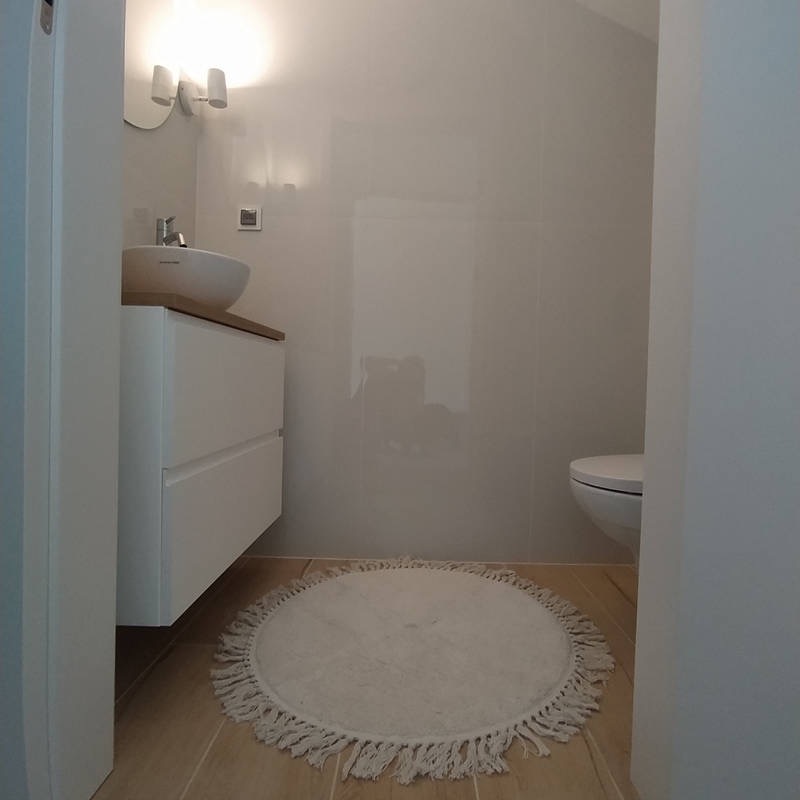 Jasna łazienka z umywalką nablatową, białymi płytkami na ścianach i okrągłym dywanikiem z frędzlami na podłodze z imitacji drewna.