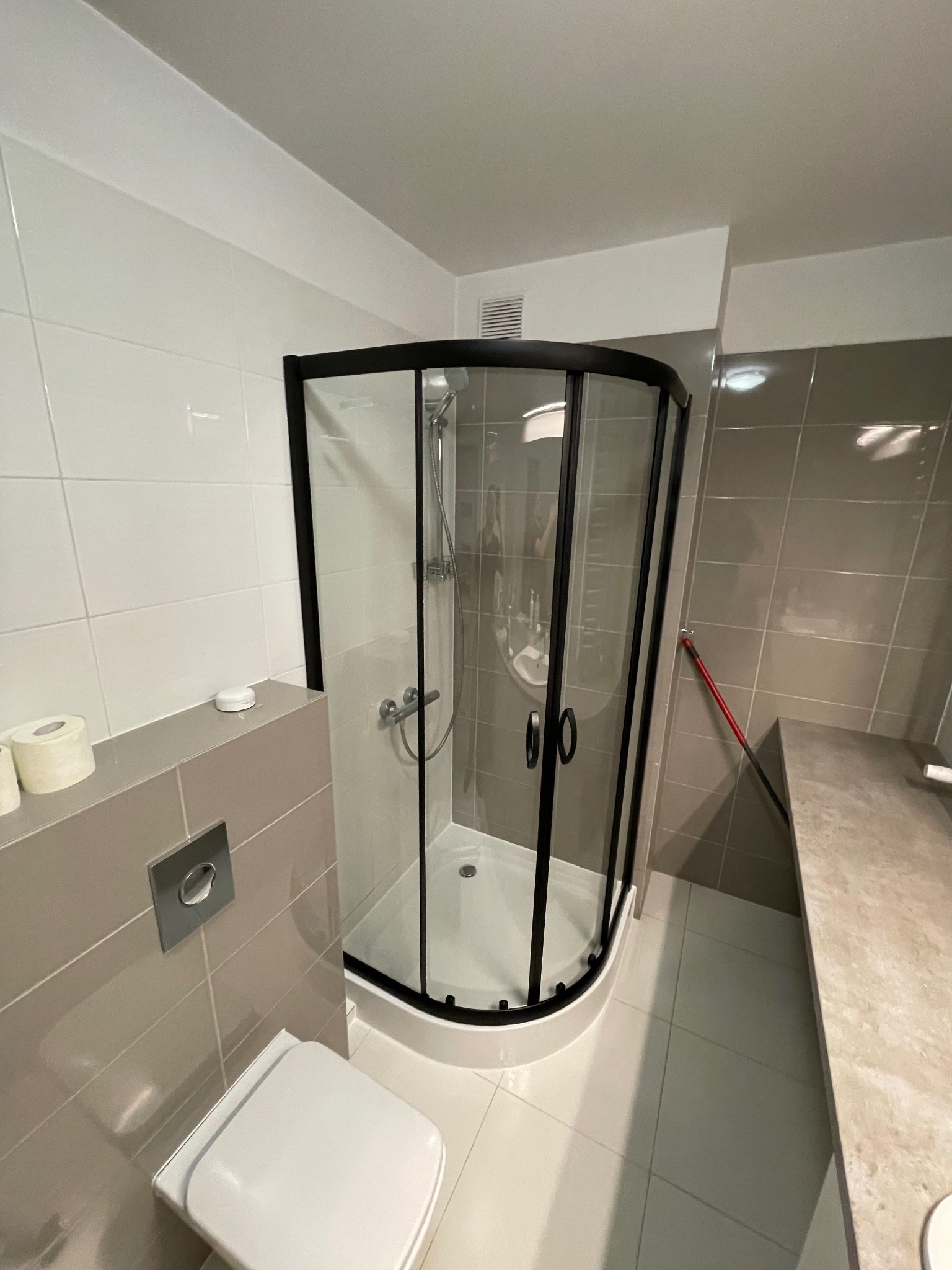 Nowoczesna łazienka z kabiną prysznicową typu walk-in z czarnymi profilami, białą ceramiką i szarymi płytkami, widoczny fragment toalety i półka z papierem toaletowym.