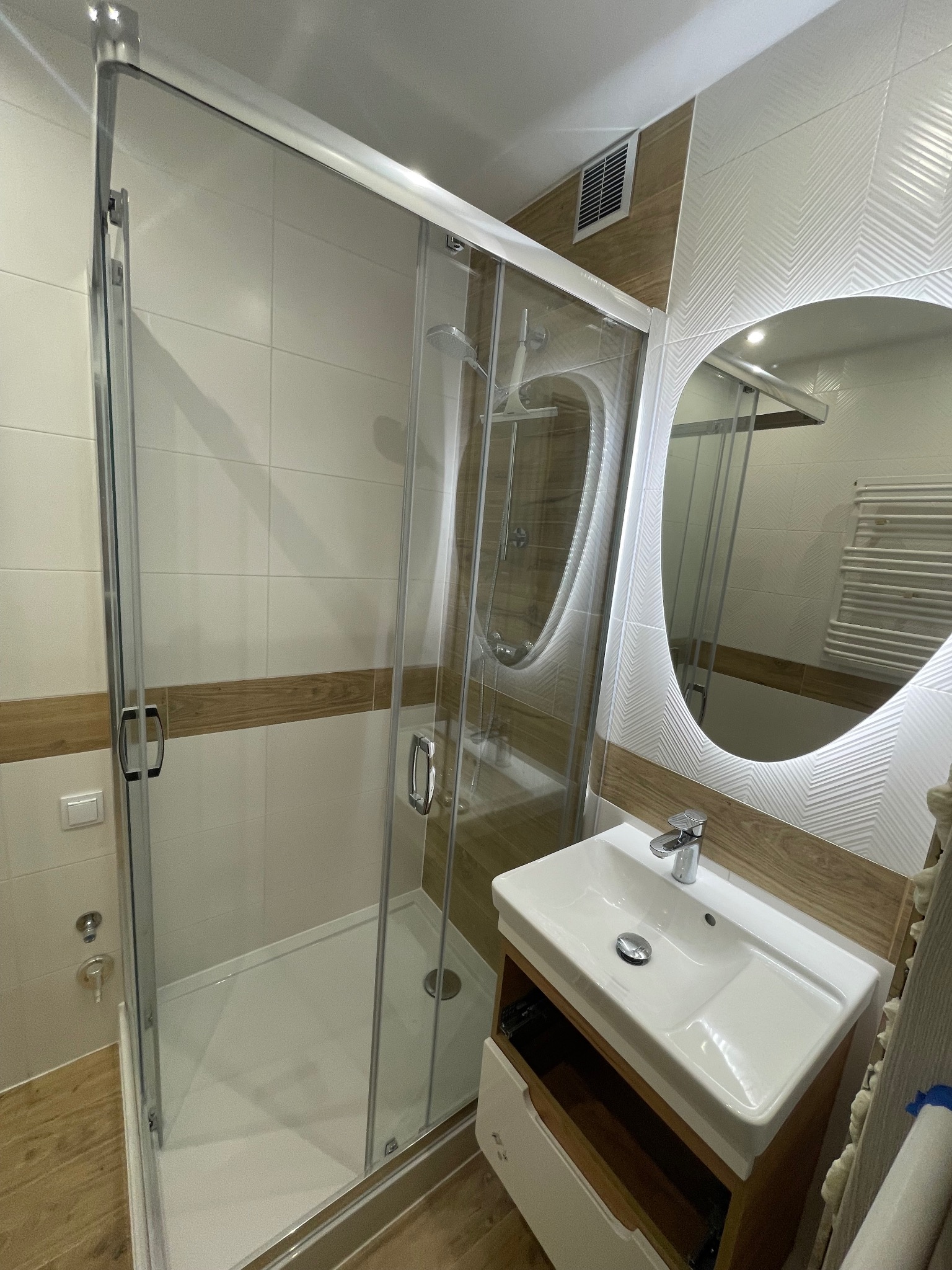 Nowoczesna łazienka z kabiną prysznicową z przesuwnymi drzwiami, umywalką nablatową na drewnianej szafce i owalnym lustrem z podświetleniem LED, wykończona białymi płytkami z drewnianymi akcentami.