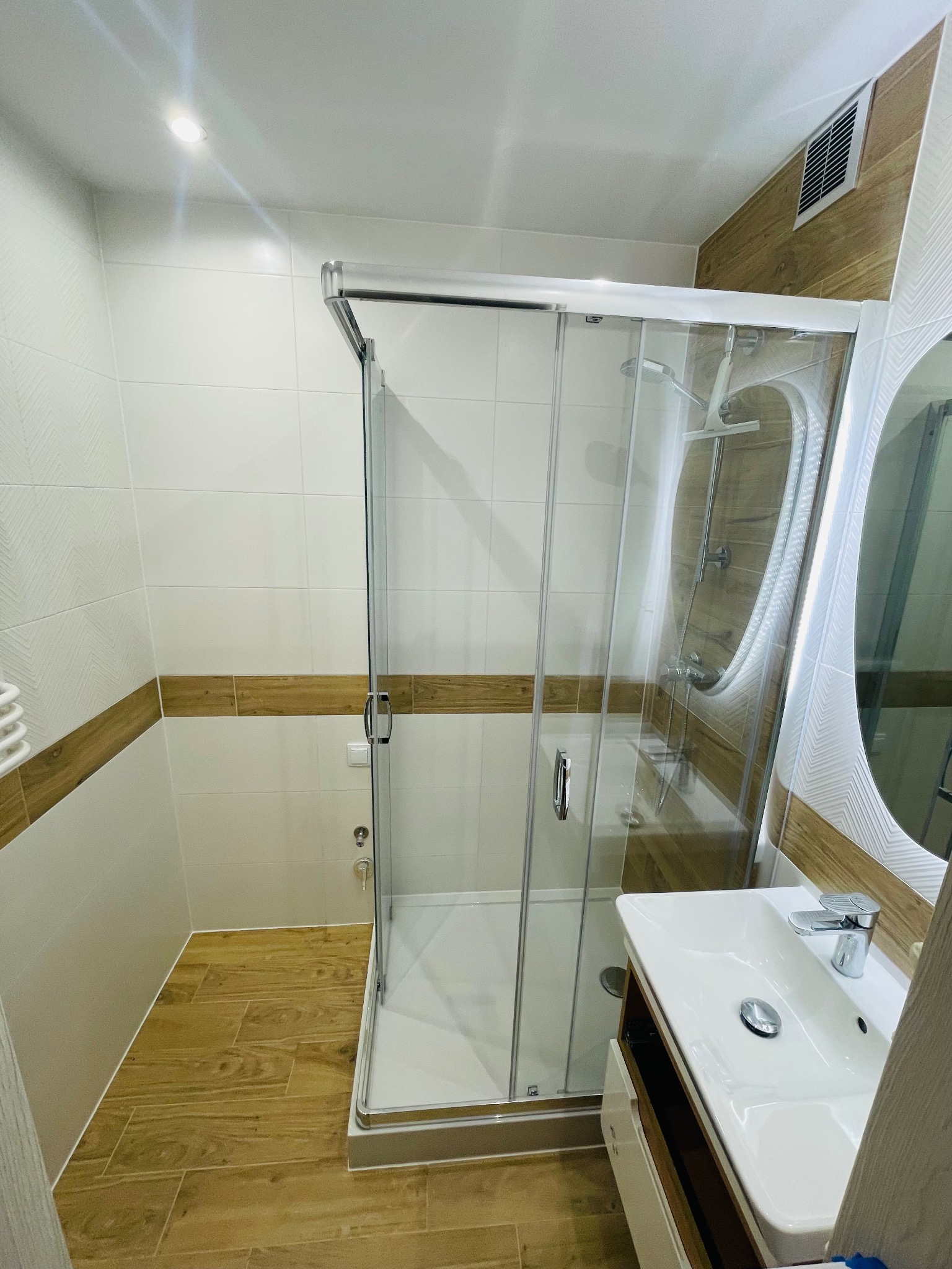 Nowoczesna łazienka po remoncie z przeszkloną kabiną prysznicową, umywalką z szafką i drewnopodobnymi akcentami na ścianach i podłodze.