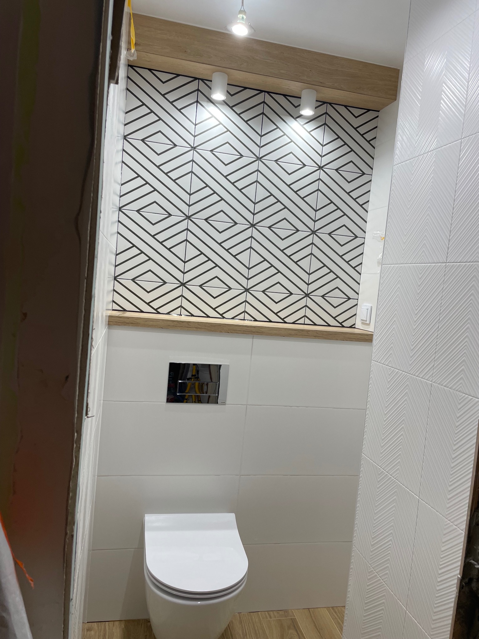 Nowoczesna toaleta podwieszana w łazience z geometryczną mozaiką na ścianie, oświetlona punktowymi lampami i wykończona drewnianym blatem.