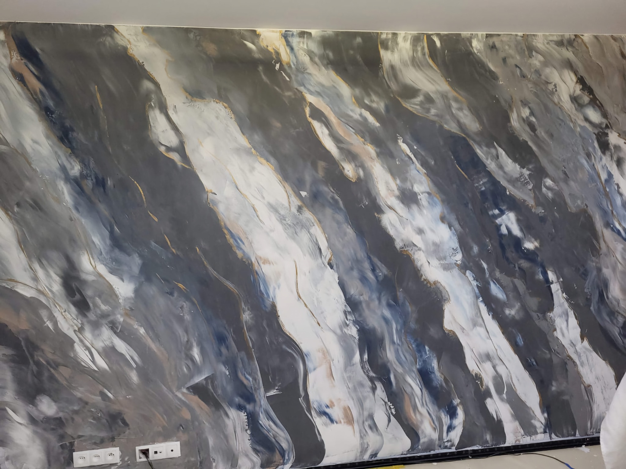 Ściana z dekoracyjną fakturą imitującą marmur w odcieniach szarości, bieli i błękitu, z delikatnymi złotymi akcentami, widoczne gniazdka elektryczne przy podłodze.