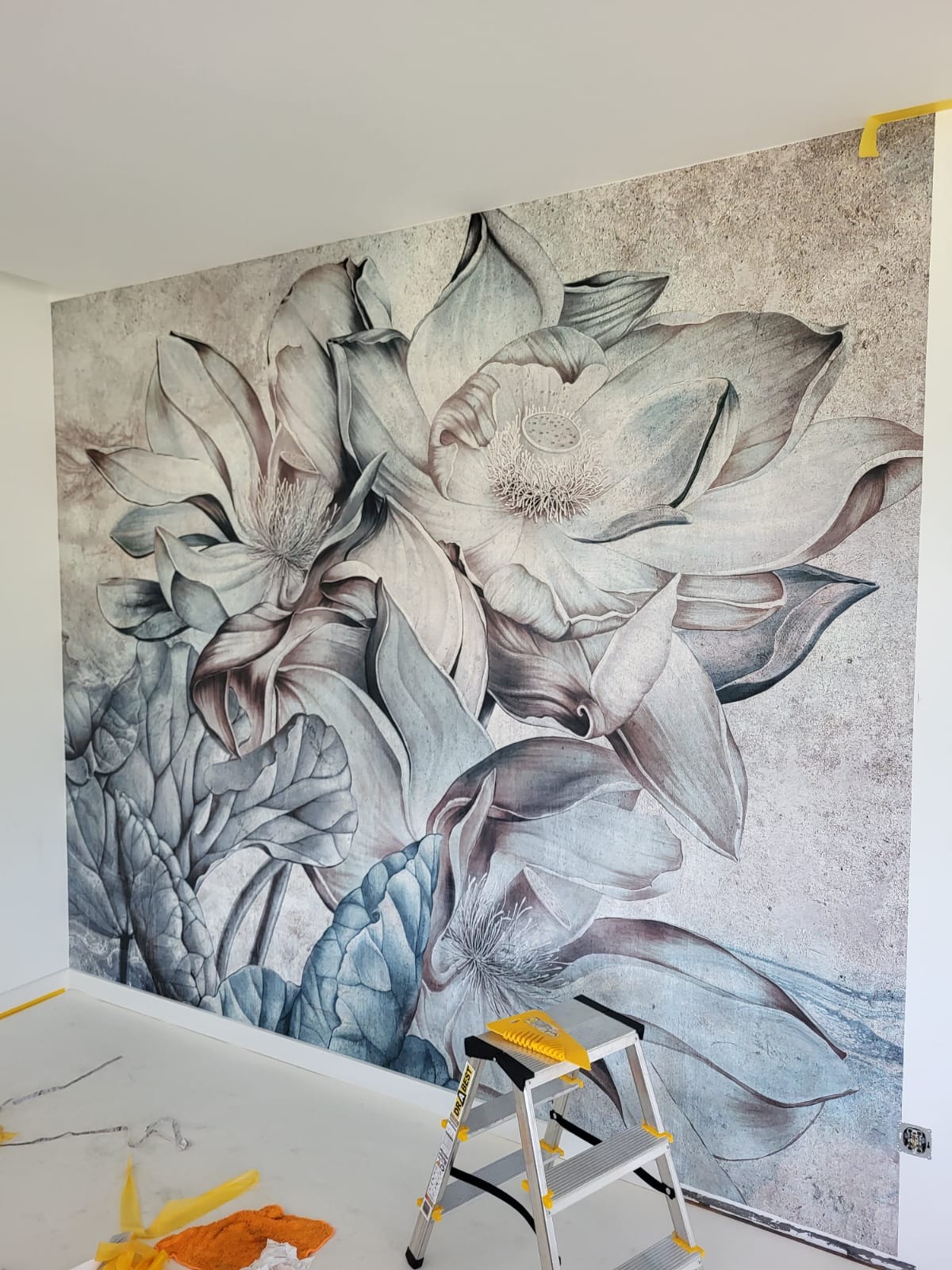 Ściana z fototapetą przedstawiającą duże kwiaty lotosu w odcieniach szarości i błękitu, widoczna drabina malarska i fragment podłogi z resztkami taśmy malarskiej.